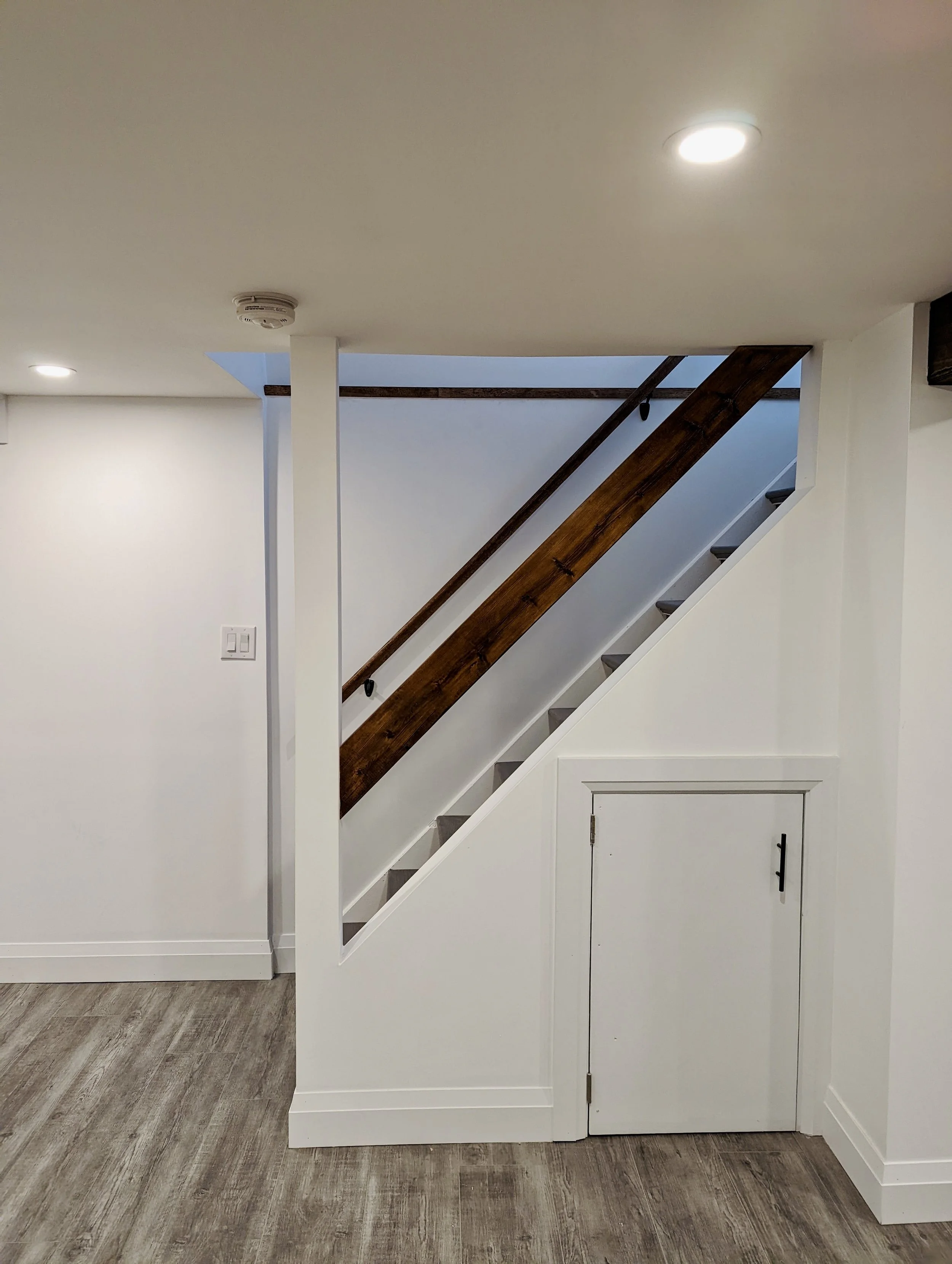 Interior view of a staircase with a wooden handrail, white walls, and a small storage door at the bottom.