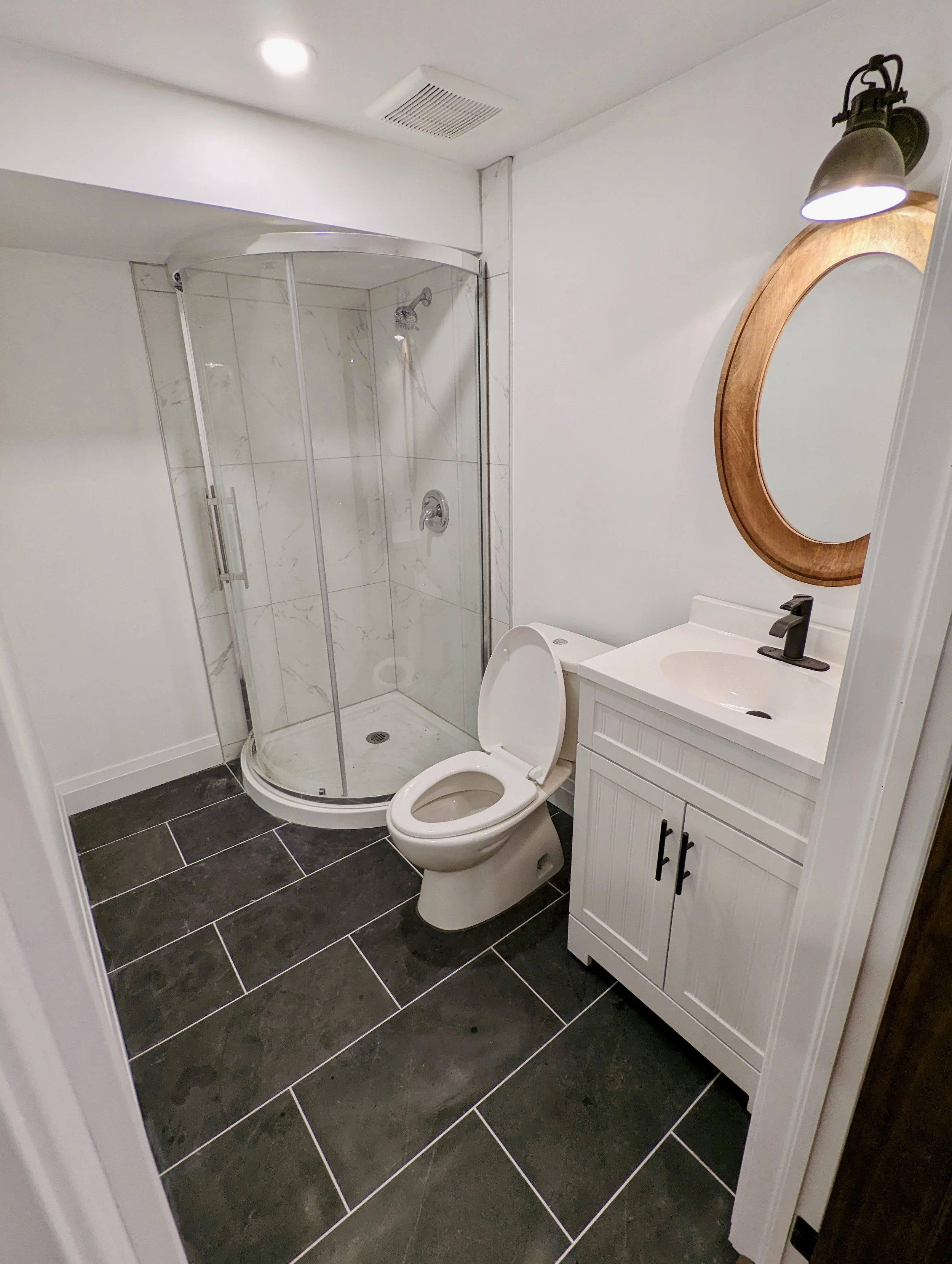 Small bathroom with black tiled floor, white vanity with a sink and oval mirror, toilet, and glass corner shower with marble tiles.