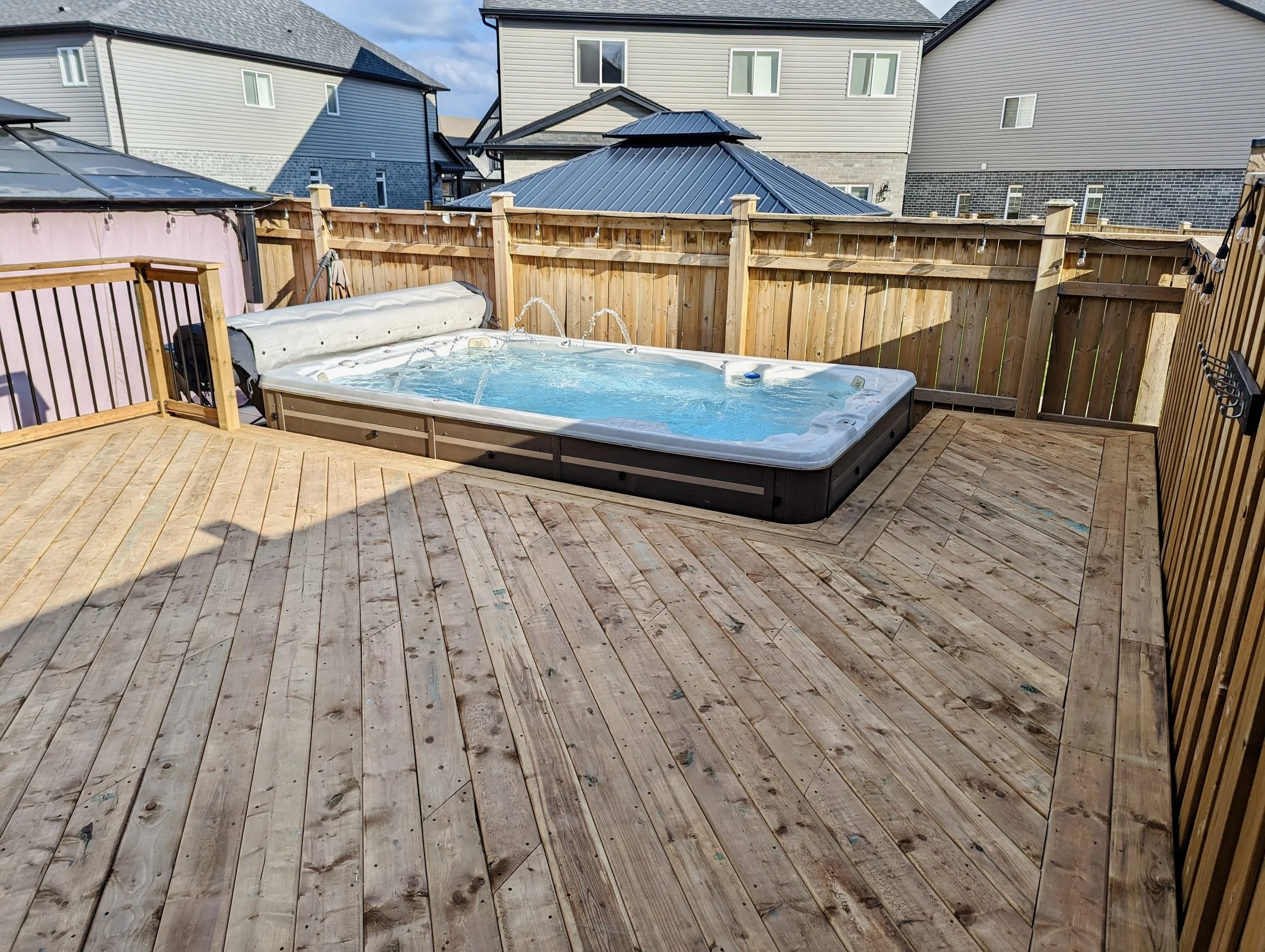 Backyard deck with a hot tub surrounded by wooden fencing and houses in the background.