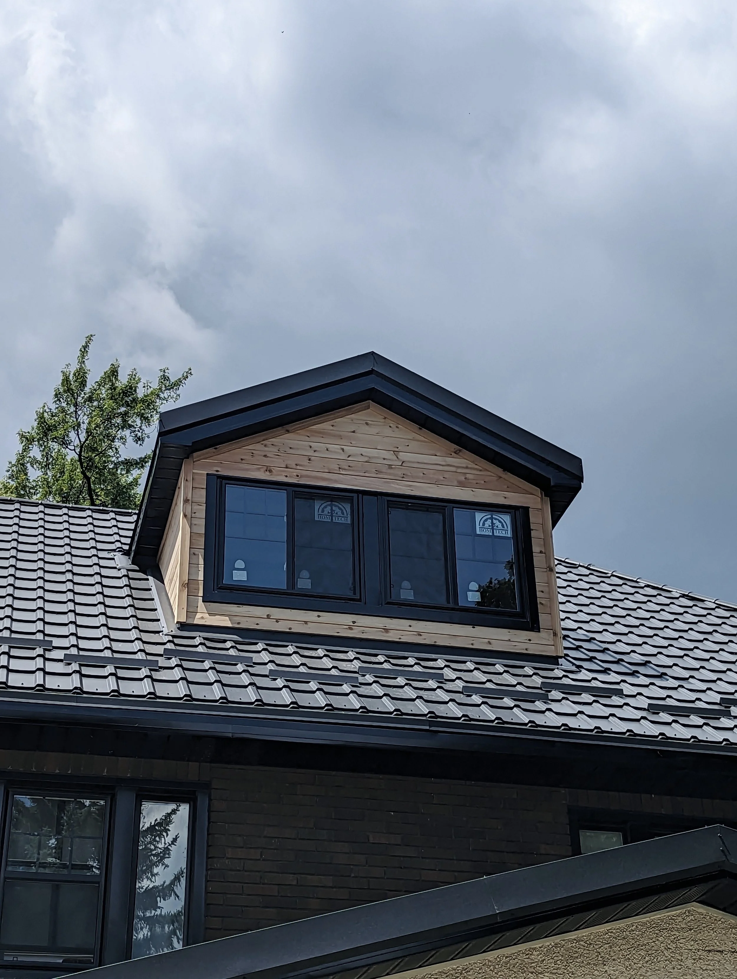 Close-up of a house dormer with black-framed windows and a black gabled roof, over a brick house with a sloped tiled roof, against a cloudy sky.