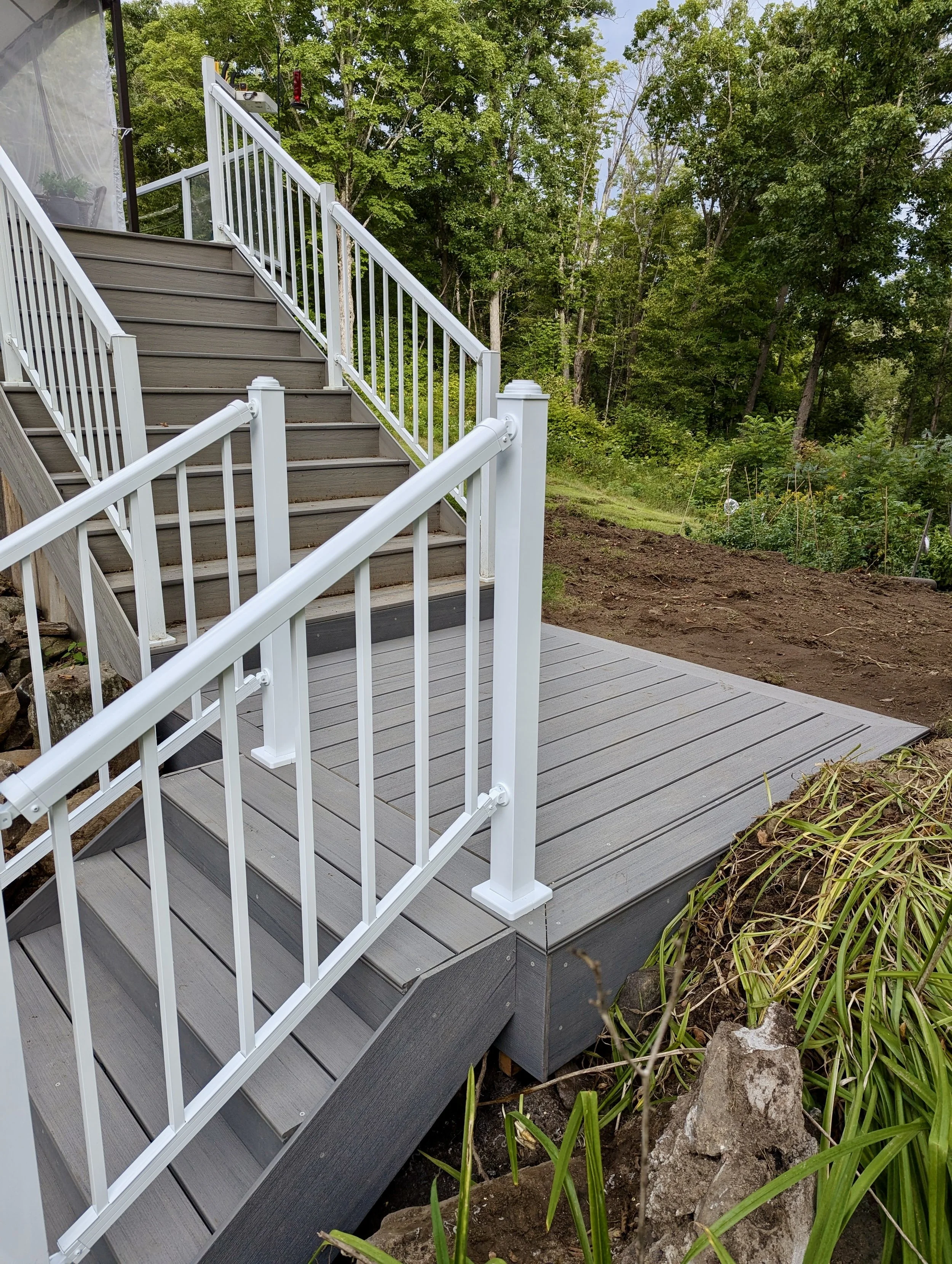 Wooden outdoor staircase and deck with white metal railings, surrounded by green trees and plants in a backyard setting.