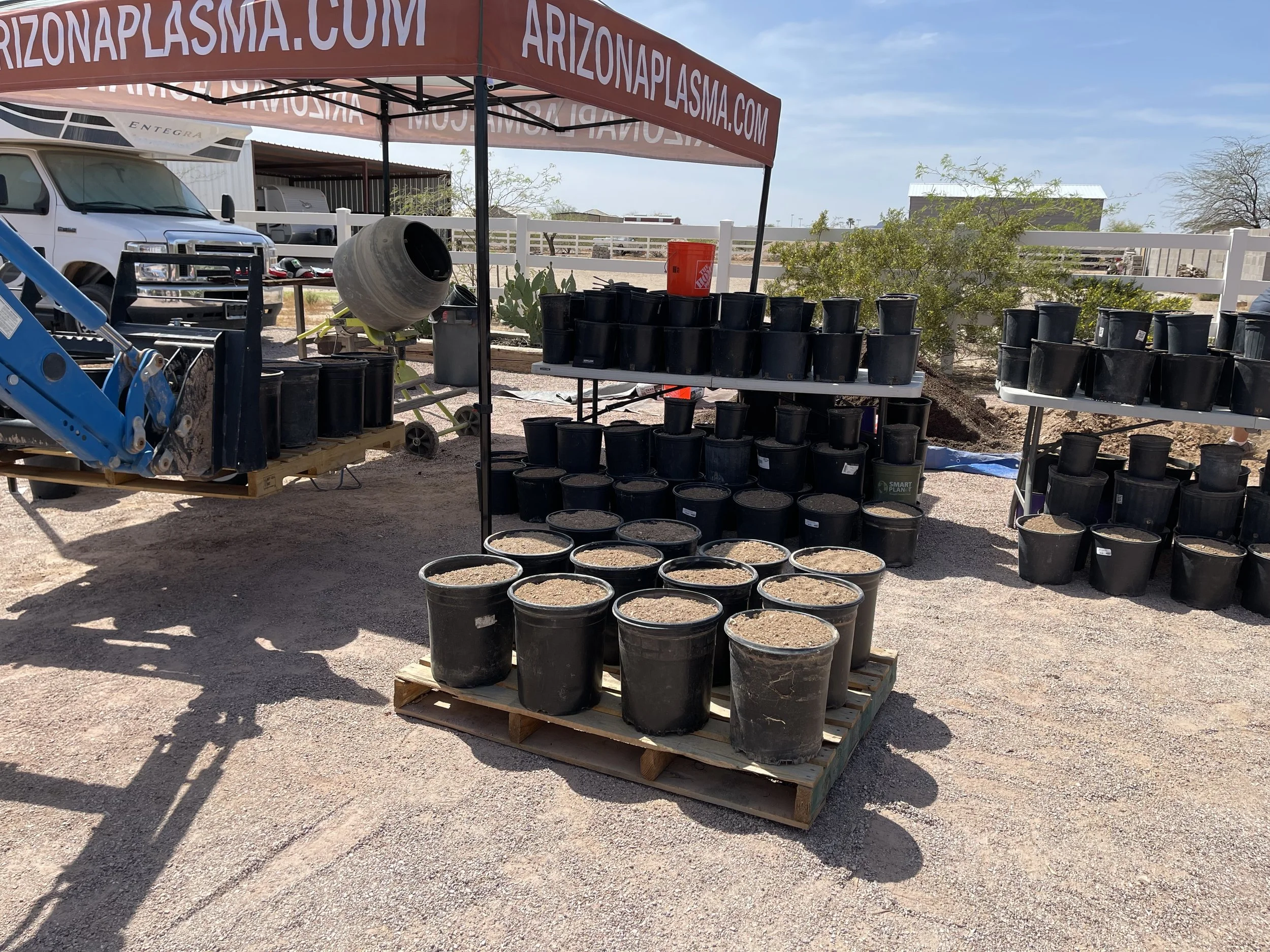 Outdoor scene with a canopy advertising 'arizonaplasma.com', metal shelves with black plant pots, some filled with dirt, on a pallet, and surrounding equipment and vehicles in a desert setting.