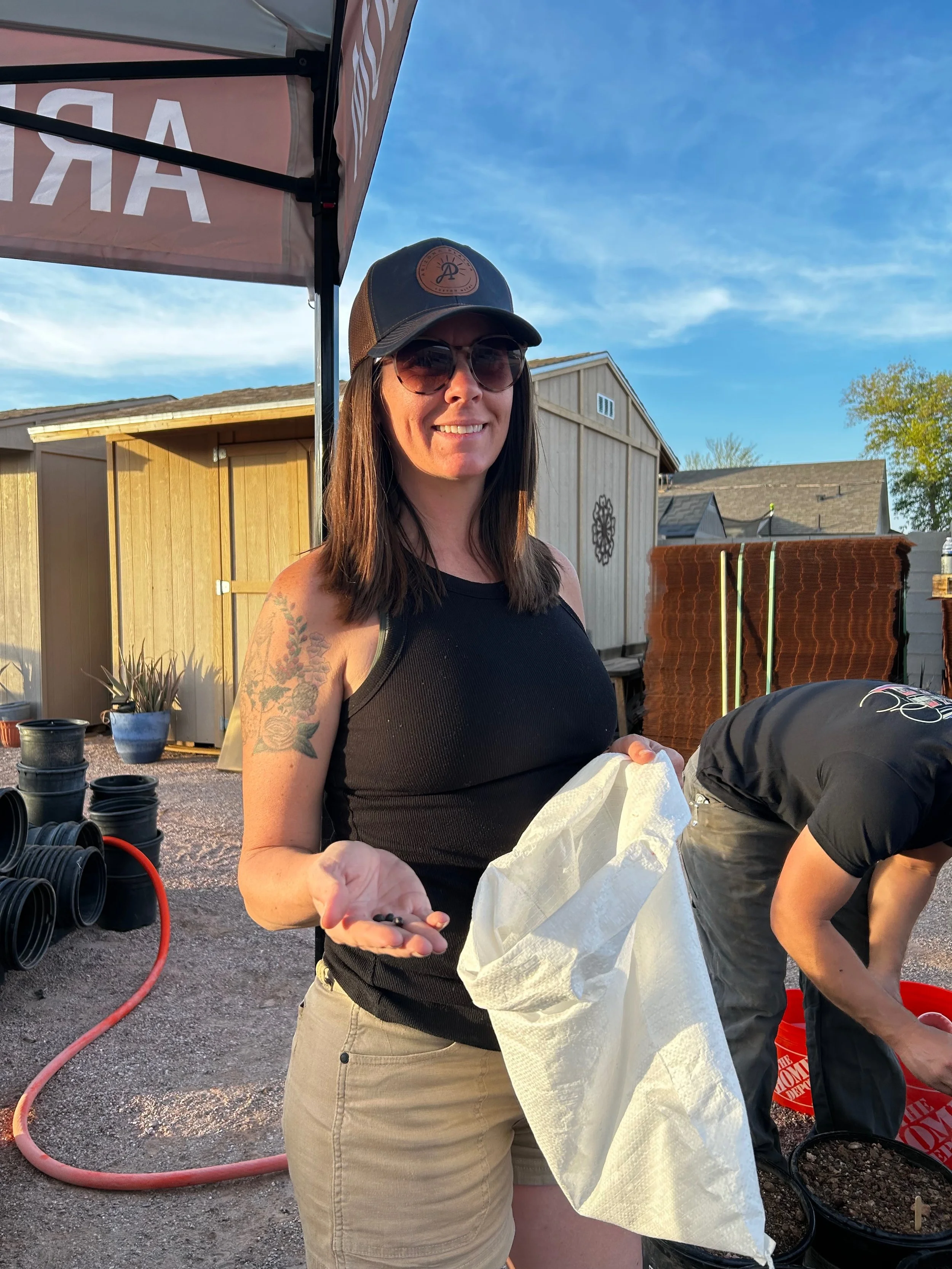 Woman with dark hair, sunglasses, and a baseball cap, smiling and holding small black objects in her hand, outdoors at a gardening or farming event.