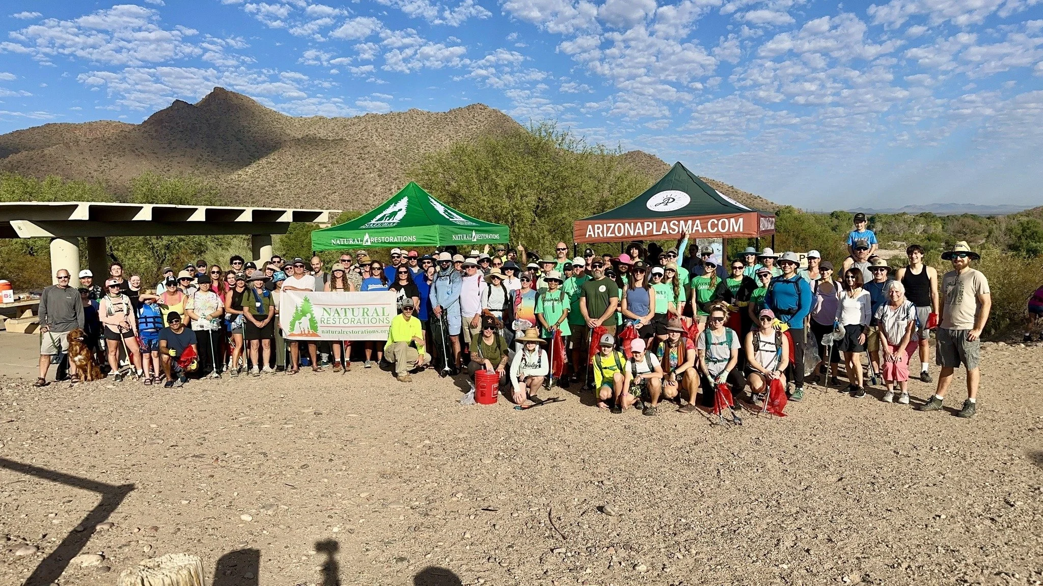 Group photo of hikers gathered outdoors near mountains, with tents displaying banners for Natural Restorations and Arizona Plasma, under a partly cloudy sky.