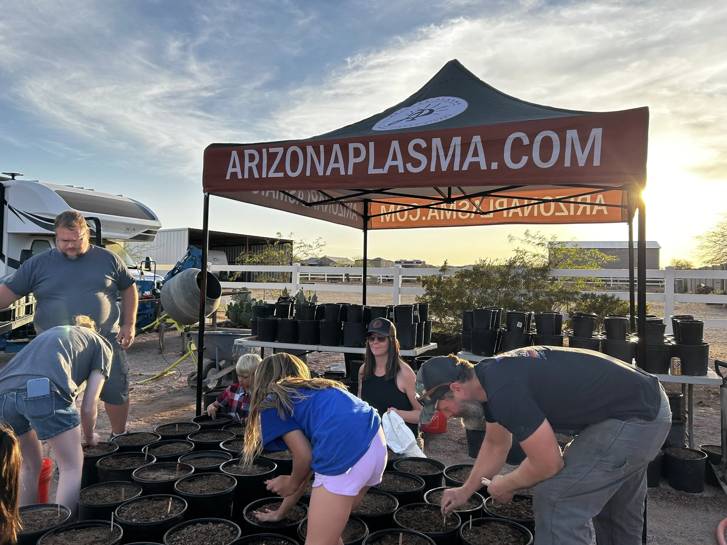 Group of people planting seedlings in black pots under a red canopy with 'ARIZONAPLASMA.COM' written on it, during sunset at outdoor farm or nursery.