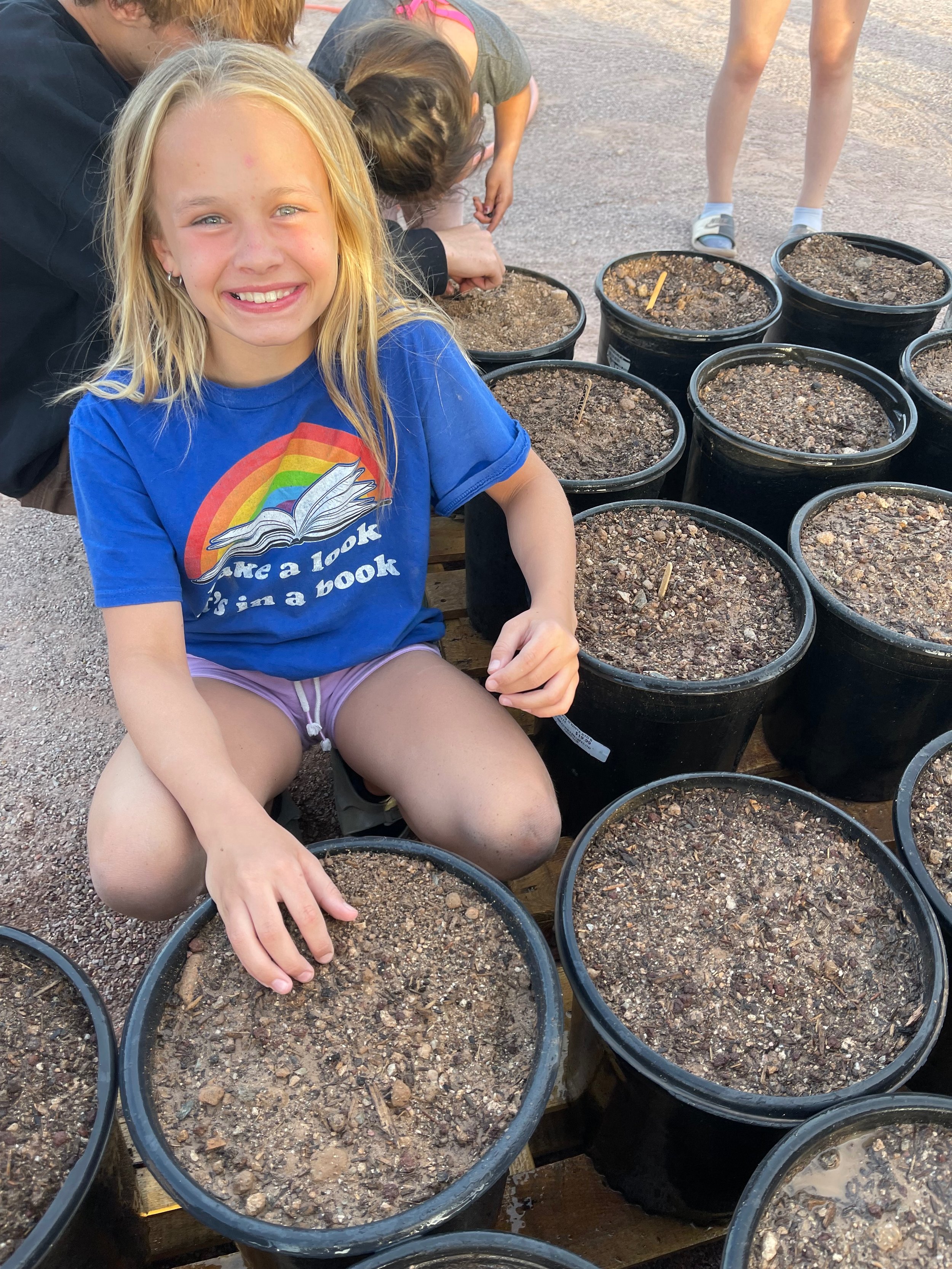 A smiling girl with long blonde hair sitting in front of multiple black plant pots filled with soil, outdoors on a sunny day.