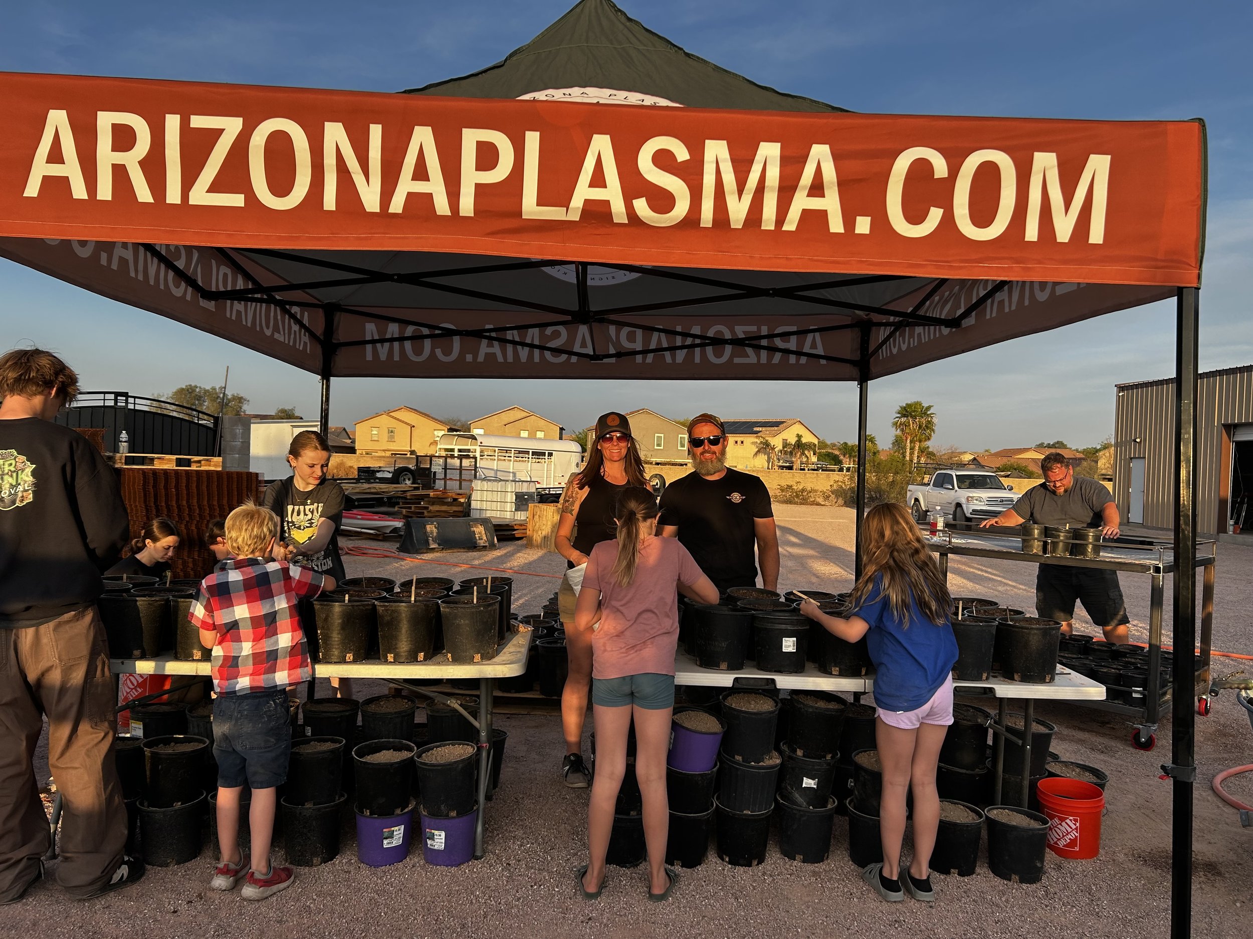 People at an outdoor booth with a large orange canopy displaying 'ARIZONAPLASMA.COM'. The booth has black buckets and supplies, and there are children and adults engaging and smiling, with houses and vehicles in the background during sunset.
