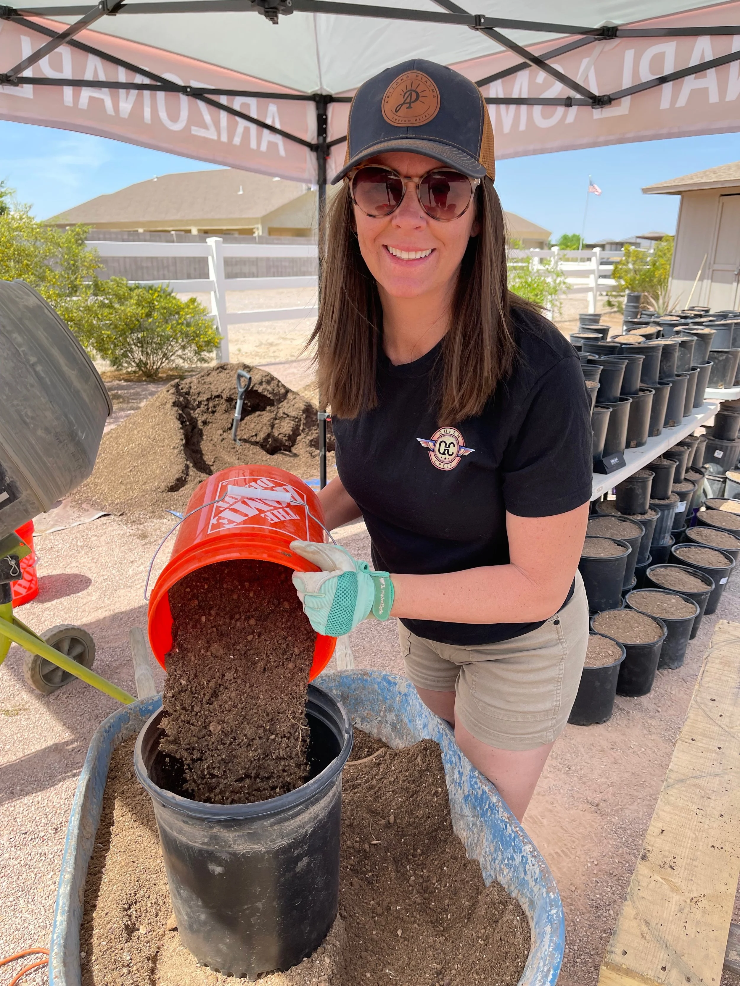 A woman with long brown hair, sunglasses, a baseball cap, a black t-shirt, and shorts is pouring soil from an orange Home Depot bucket into a black plant pot under a canopy tent, with multiple similar plant pots in the background.