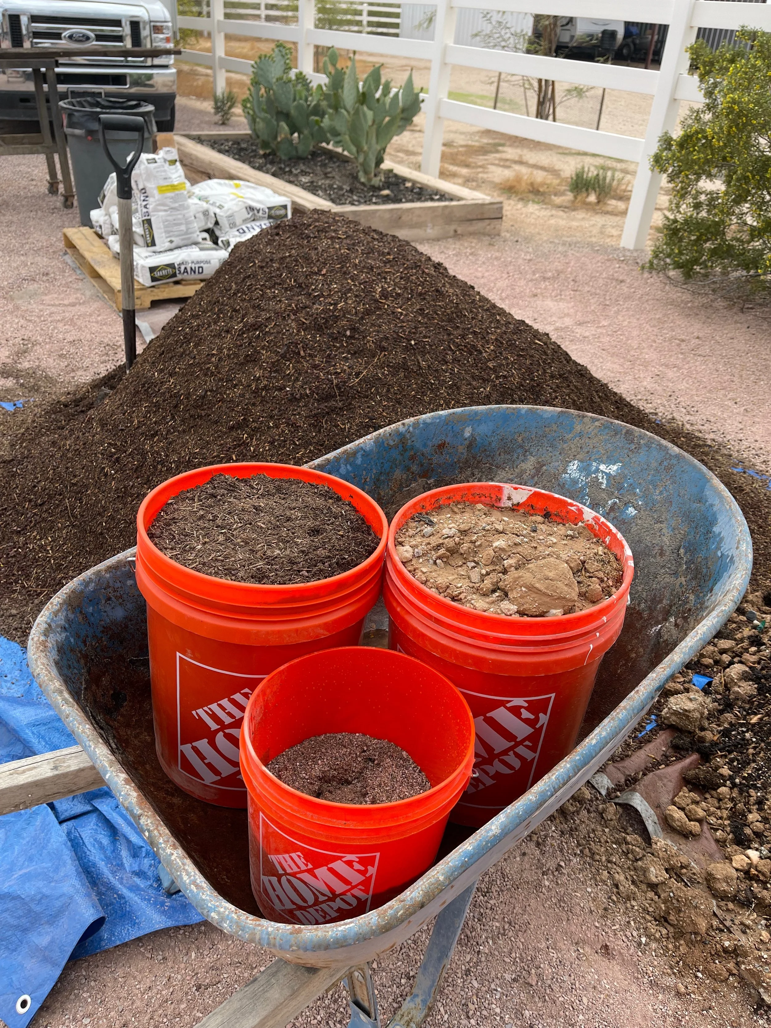 A wheelbarrow containing three orange buckets filled with different types of soil and compost, placed on a wooden construction stand outdoors near a garden bed with cactus and plants, against a background of a white fence and gravel ground.