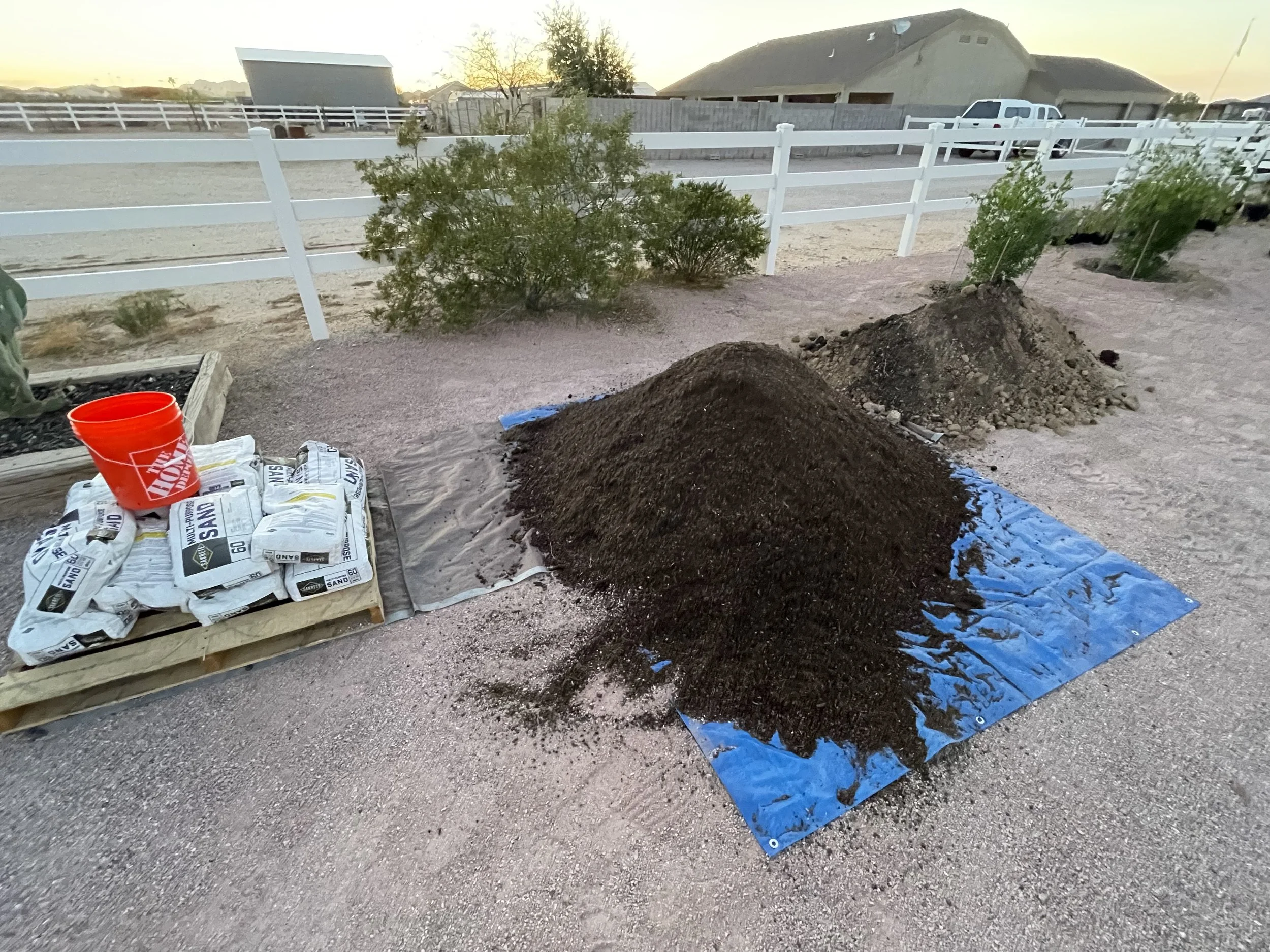 Three piles of soil or compost on a blue tarp outside, with gardening supplies and a red bucket nearby, in a yard enclosed by a white fence at sunset.