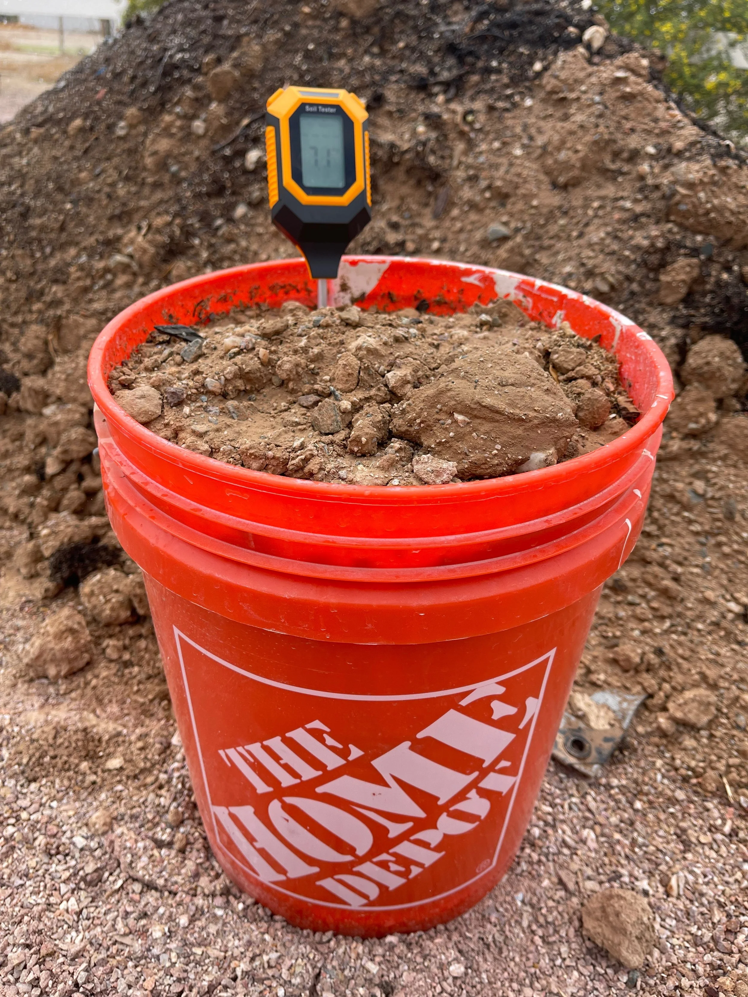 A red Home Depot bucket filled with soil, with a soil tester inserted into it showing a moisture level of 77, placed on dirt at a construction site.
