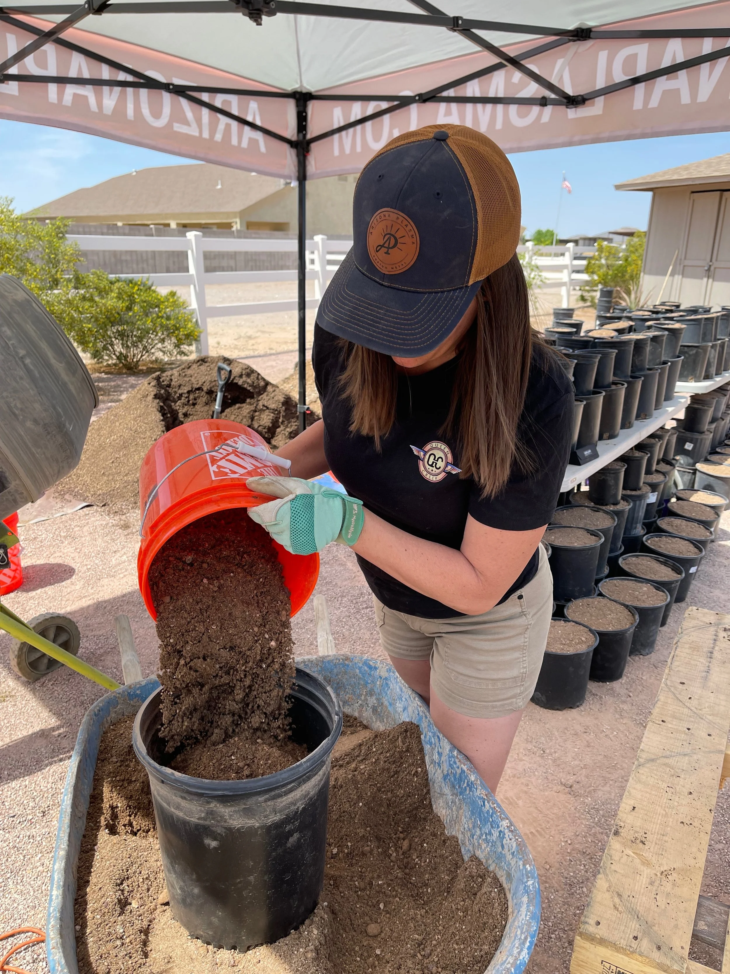 A woman wearing a black cap, black t-shirt, and khaki shorts, using her hands with gloves to pour soil from an orange bucket into a black pot, set on a wheelbarrow, outdoors under a canopy.