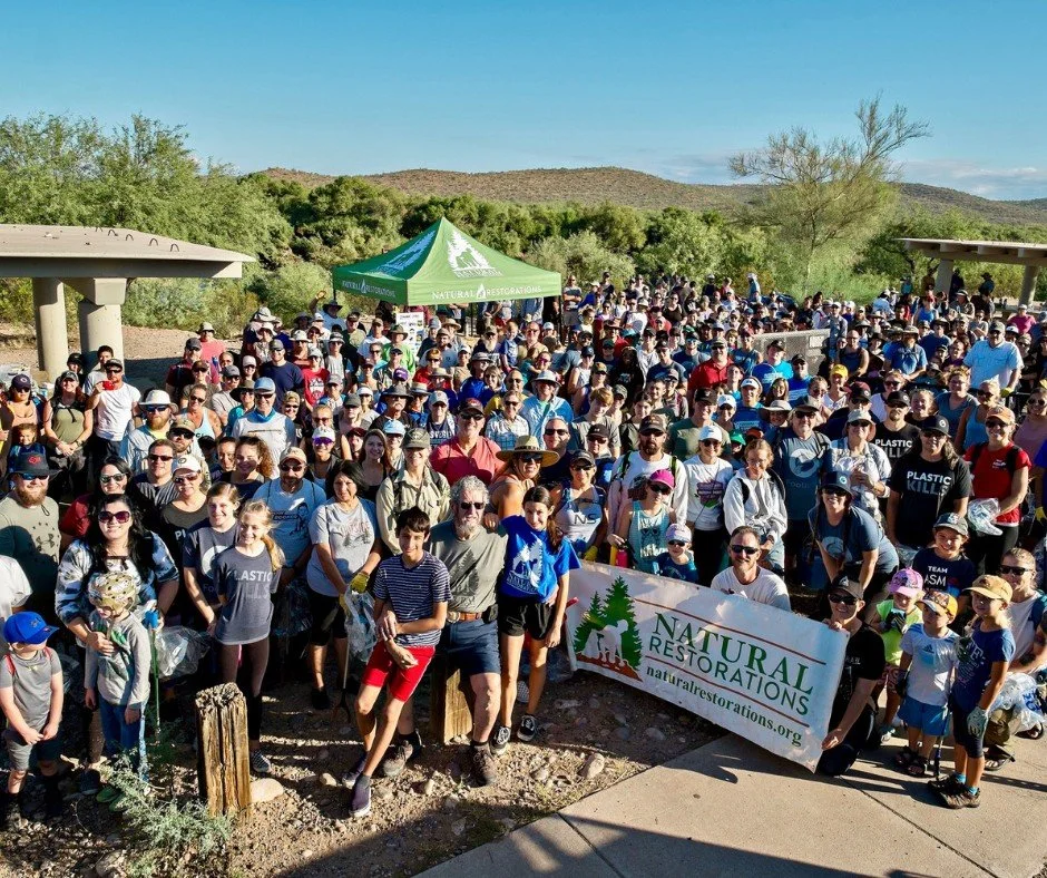 A large group of people gathered outdoors during daytime for an event organized by Natural Restorations, with a green tent and desert landscape in the background.