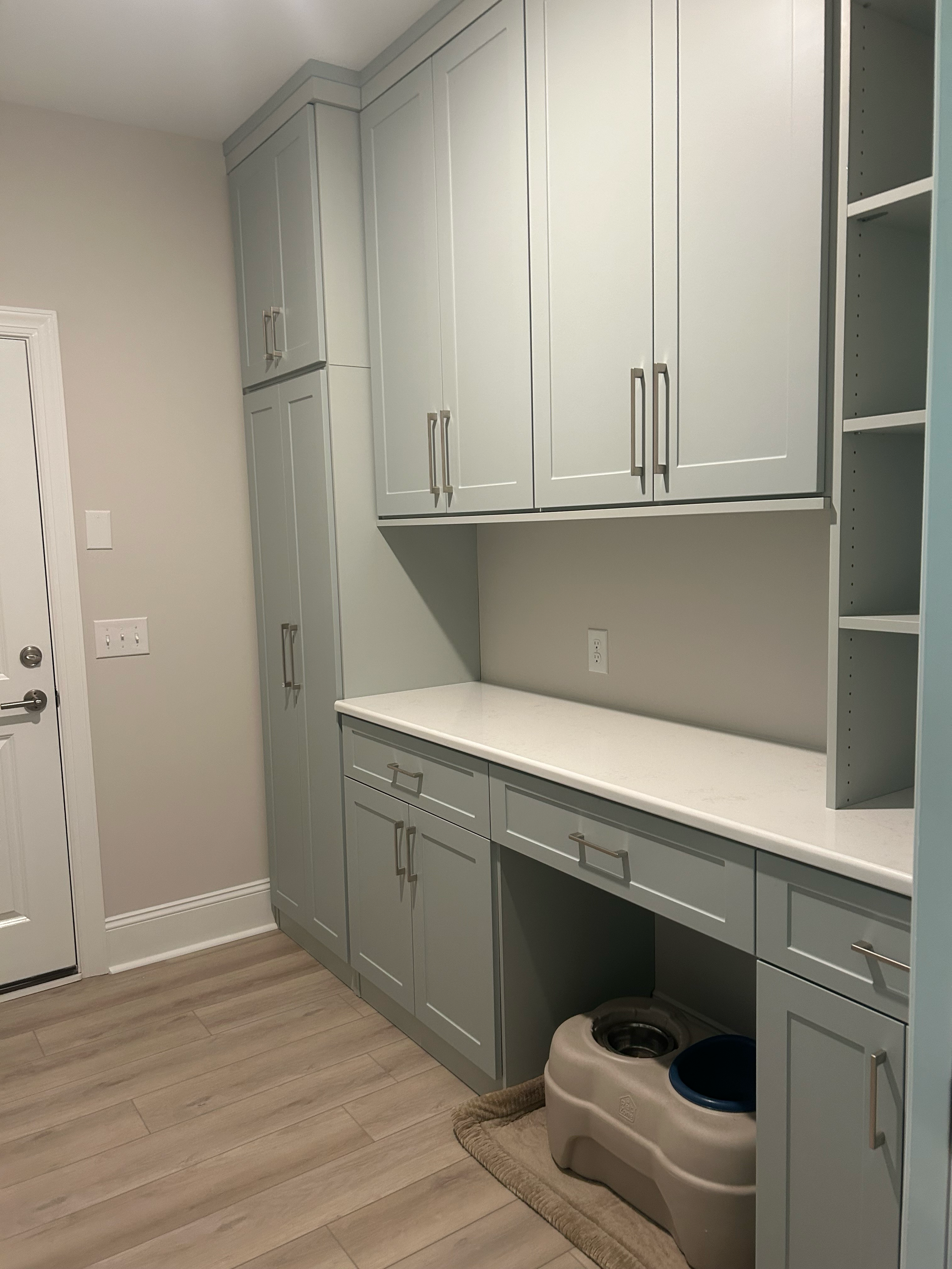 A custom laundry room storage design with light gray cabinets, a white countertop, and a pet feeding station on the floor.