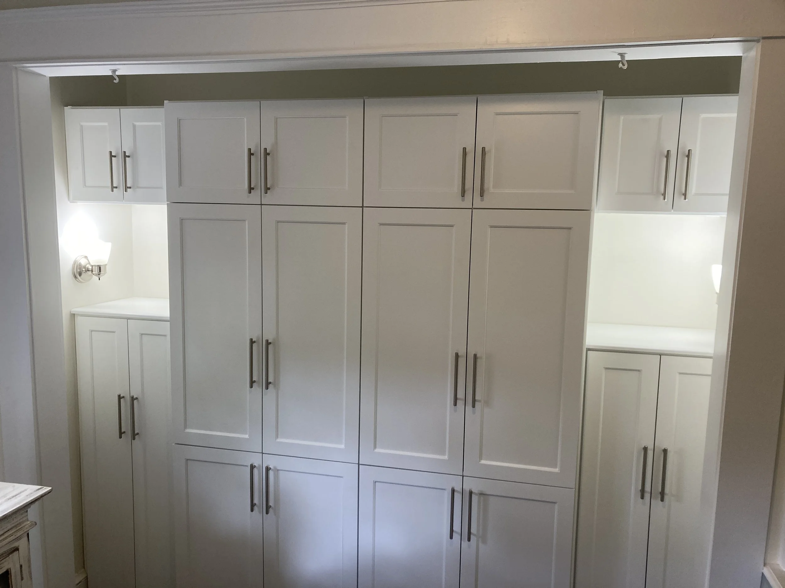 White custom built-in cabinets in a living room with silver handles in a room, two small illuminated seating areas on each side, and a wall-mounted light fixture on the left.