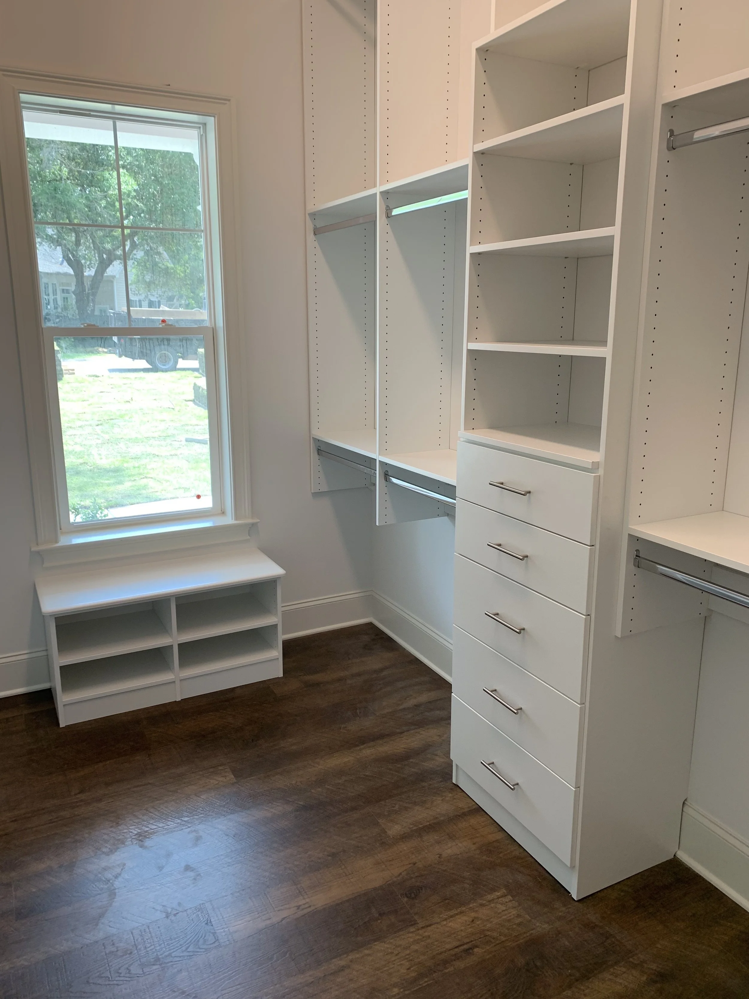 Walk-in closet with white custom-built-in shelves, drawers, and hanging rods, next to a window with a view of a green yard and a house outside.