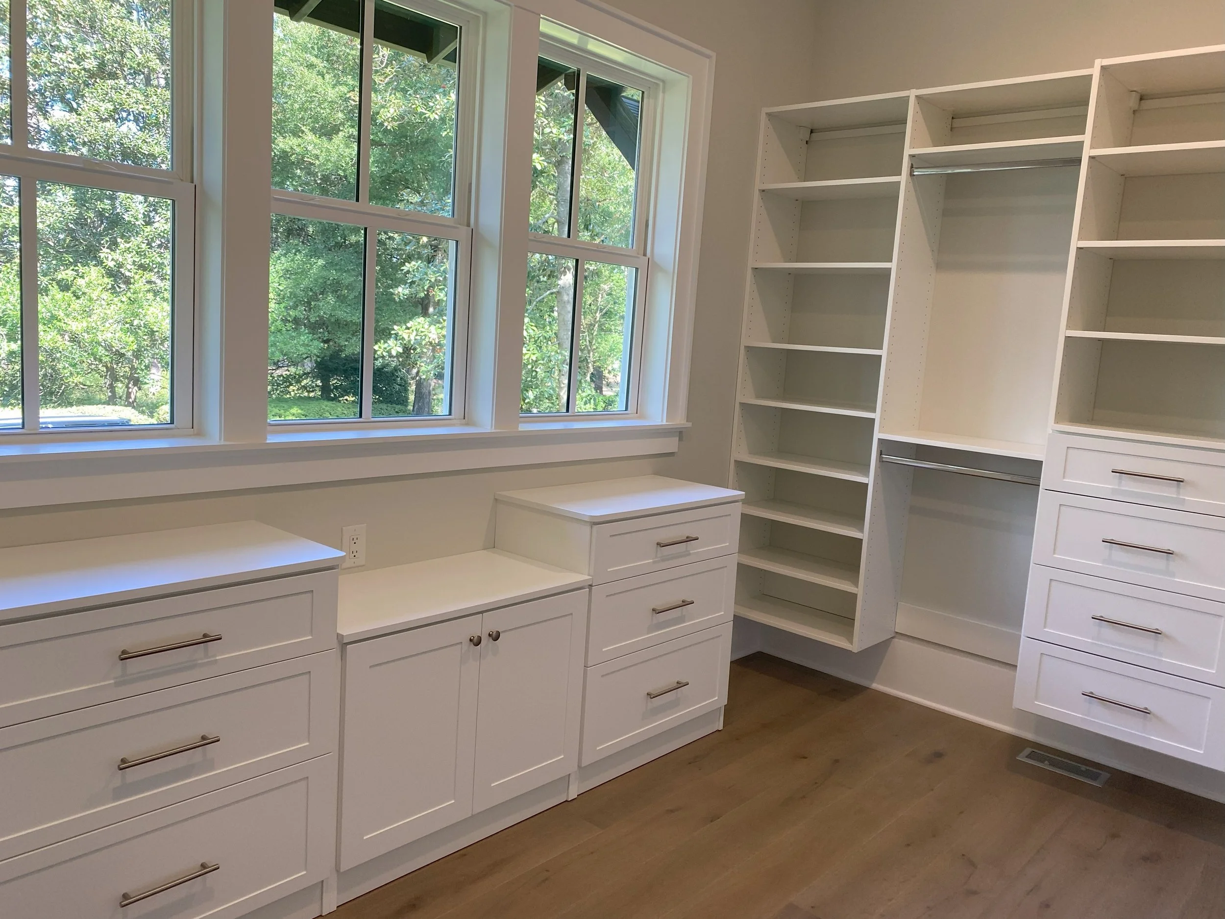 Empty custom walk-in closet with white built-in shelves and drawers, large windows with a view of green trees outside, and a hardwood floor.