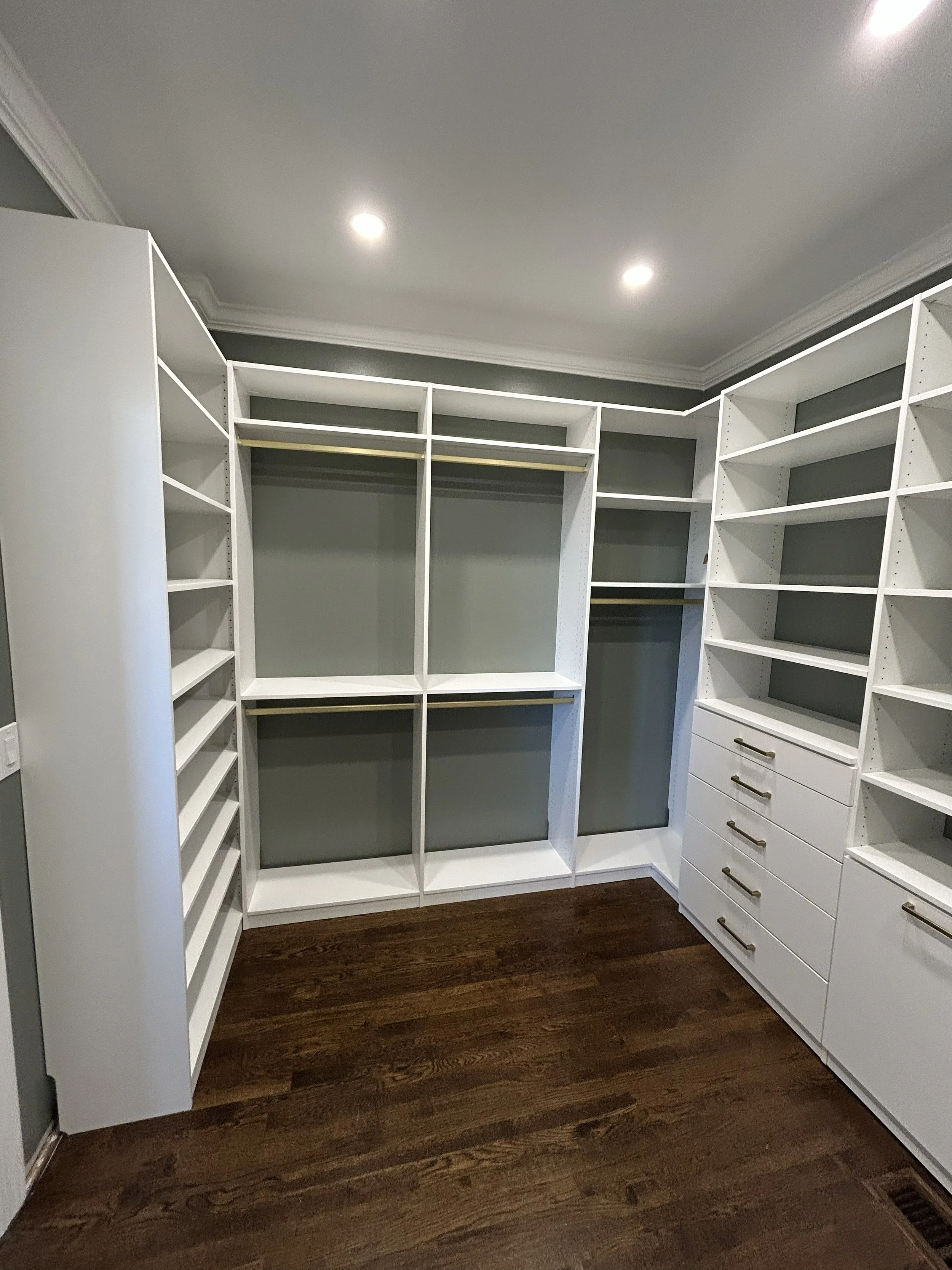 Walk-in master closet design with white built-in shelves, drawers, and hanging rods, dark wood flooring, and recessed ceiling lights.