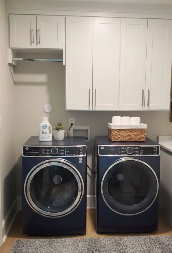 Laundry room with white cabinets, a washing machine, and a dryer. Items on top include a laundry detergent bottle, a small potted plant, and a basket of towels.