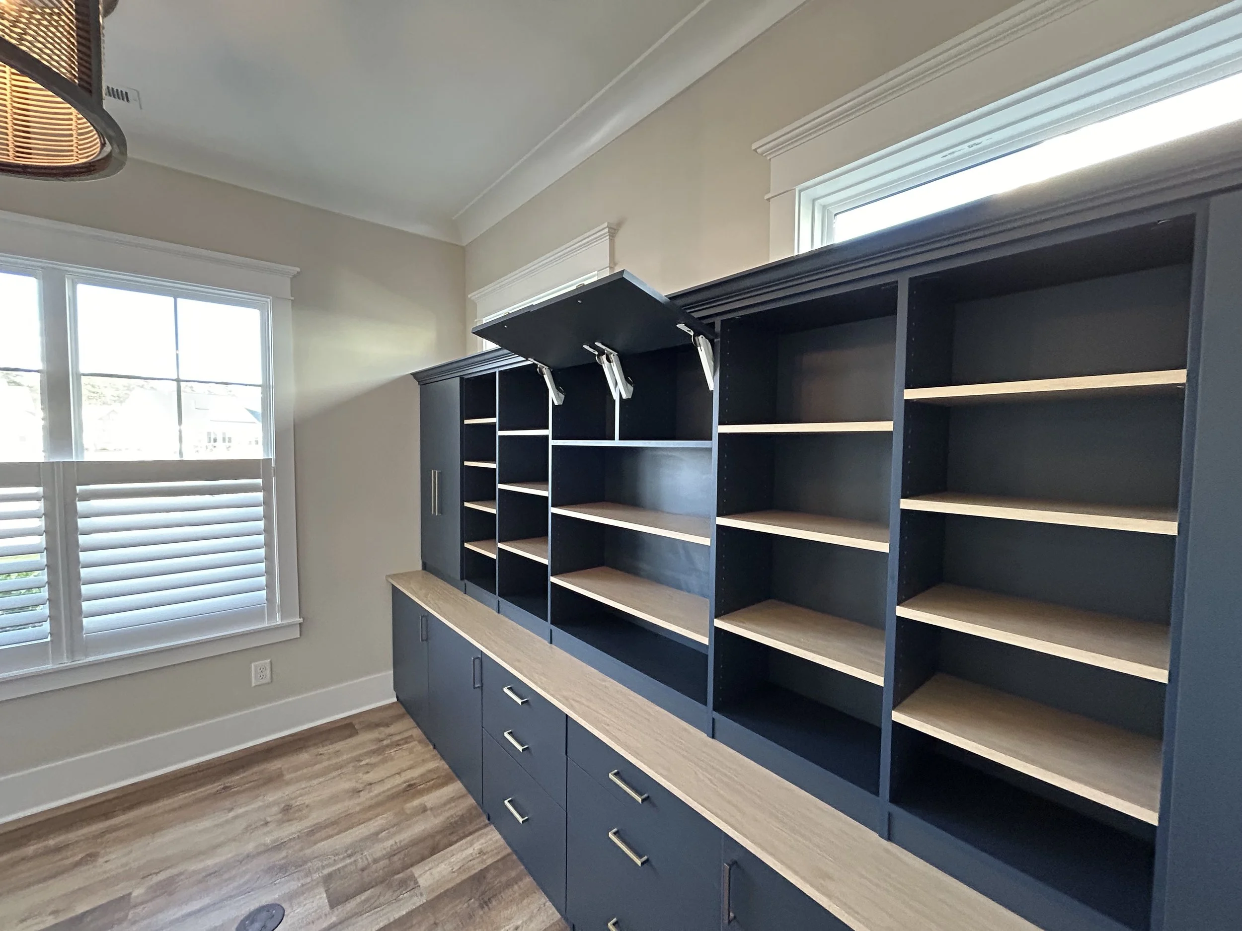 Dark blue custom bookshelf with storage cabinets with open top compartments and closed drawers beneath, located near a window in a room with light walls and wooden flooring.