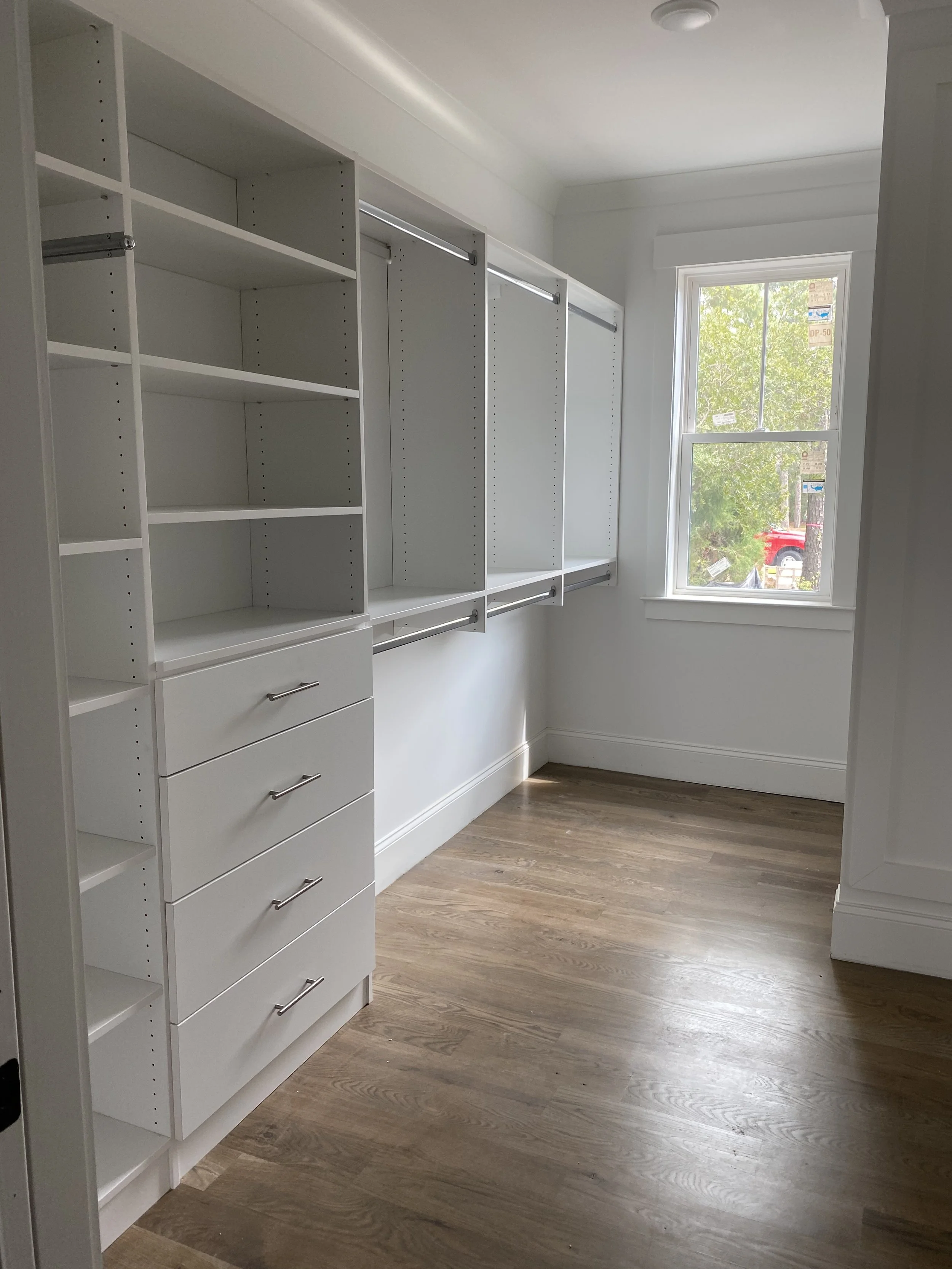 Custom closet with white shelving units and drawers, a window letting in natural light, and wooden flooring.