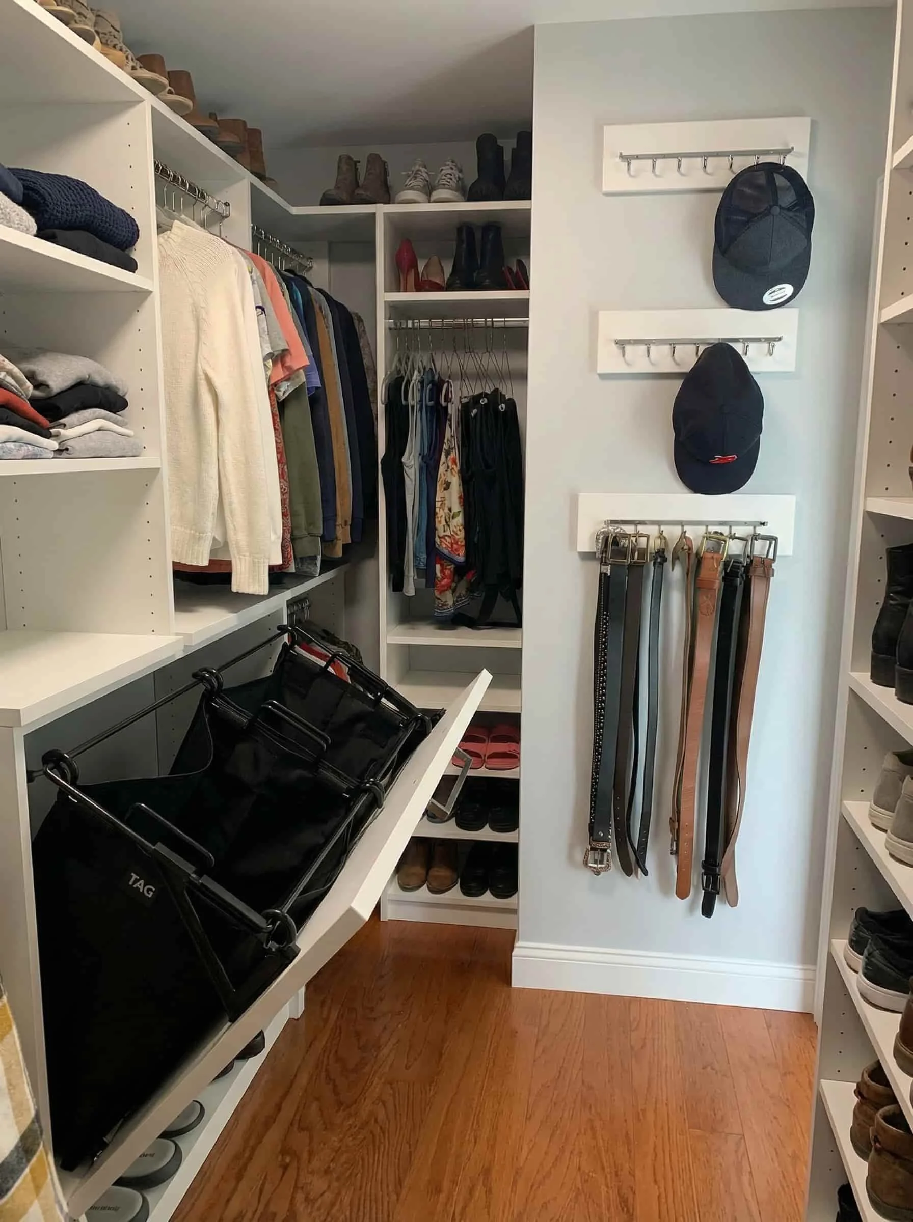 Organized walk-in closet with shelves of shoes, hanging clothes, hats, belts, and a laundry basket.