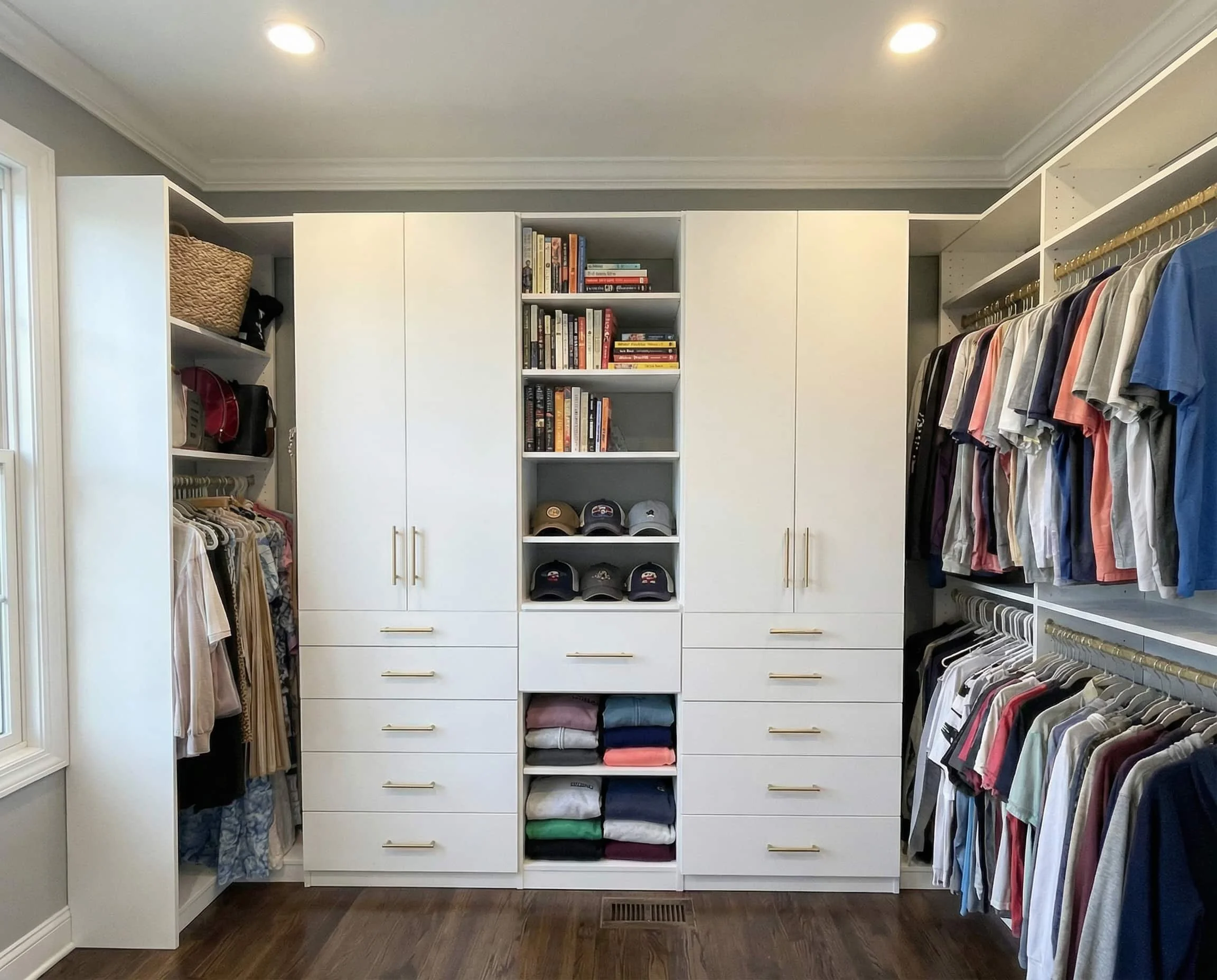 A spacious walk-in closet with white cabinets, shelves, and drawers filled with clothes, hats, books, and accessories. Hanging clothes are organized on both sides, and there is a window letting in natural light.