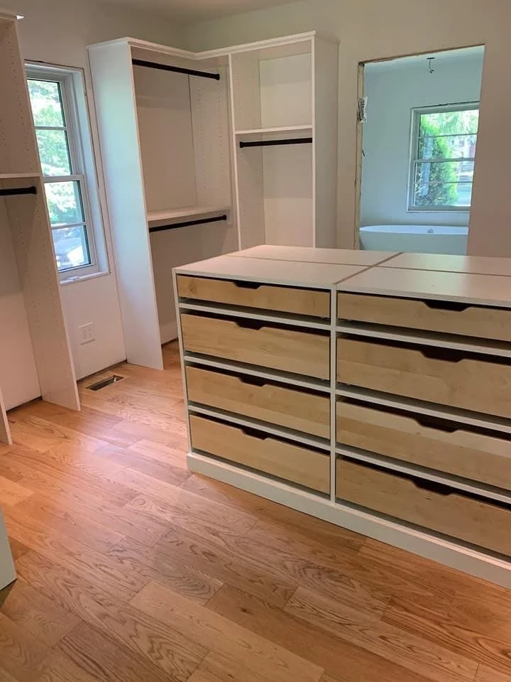Walk-in closet design with built-in shelves, drawers, and closets. Natural light from windows, hardwood floor, and an open doorway to another room with a window and bathtub visible.