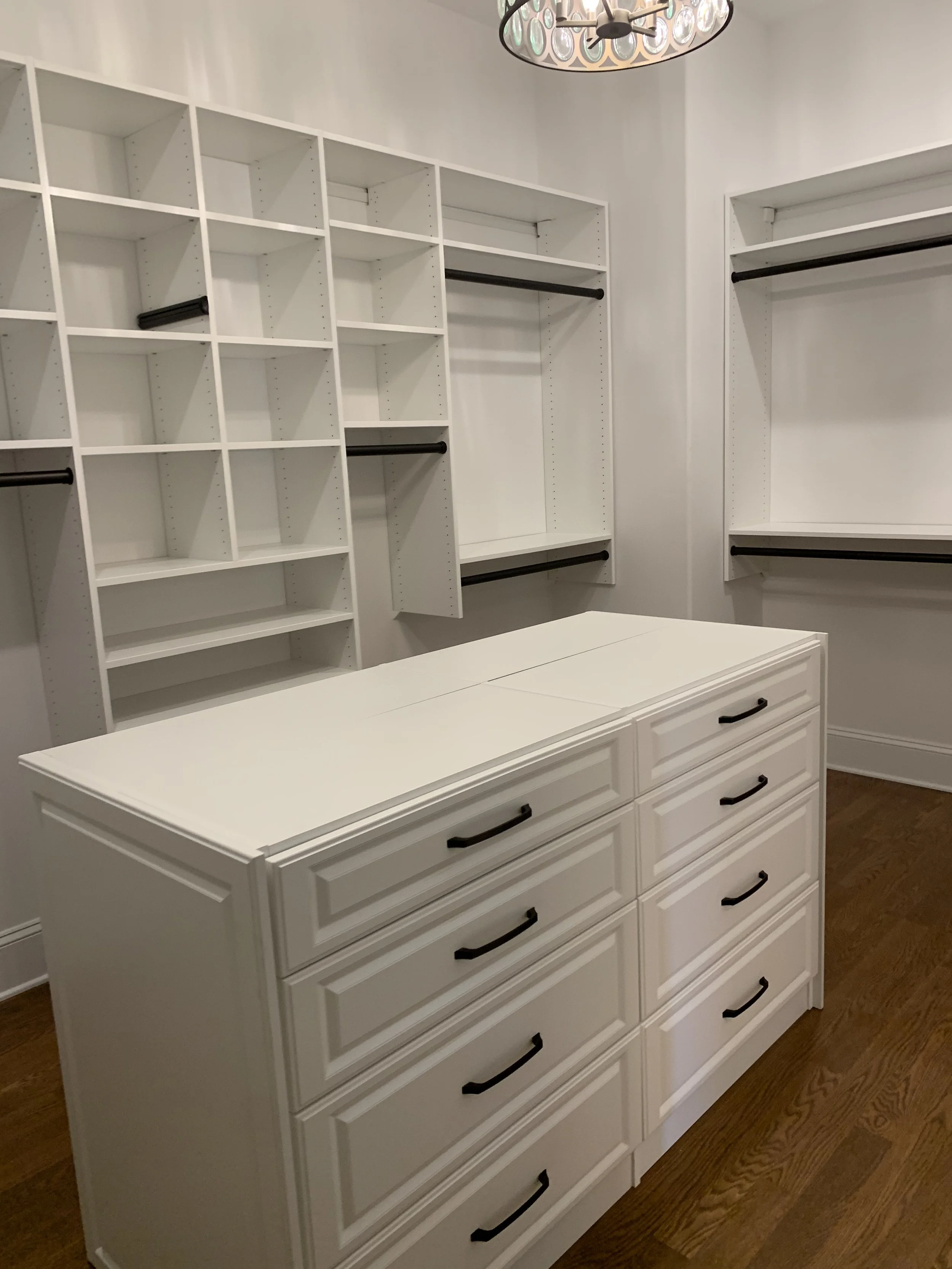 White walk-in closet with and an island surrounded b built-in shelves and drawers, and black handles, hardwood floor, and a chandelier ceiling light.