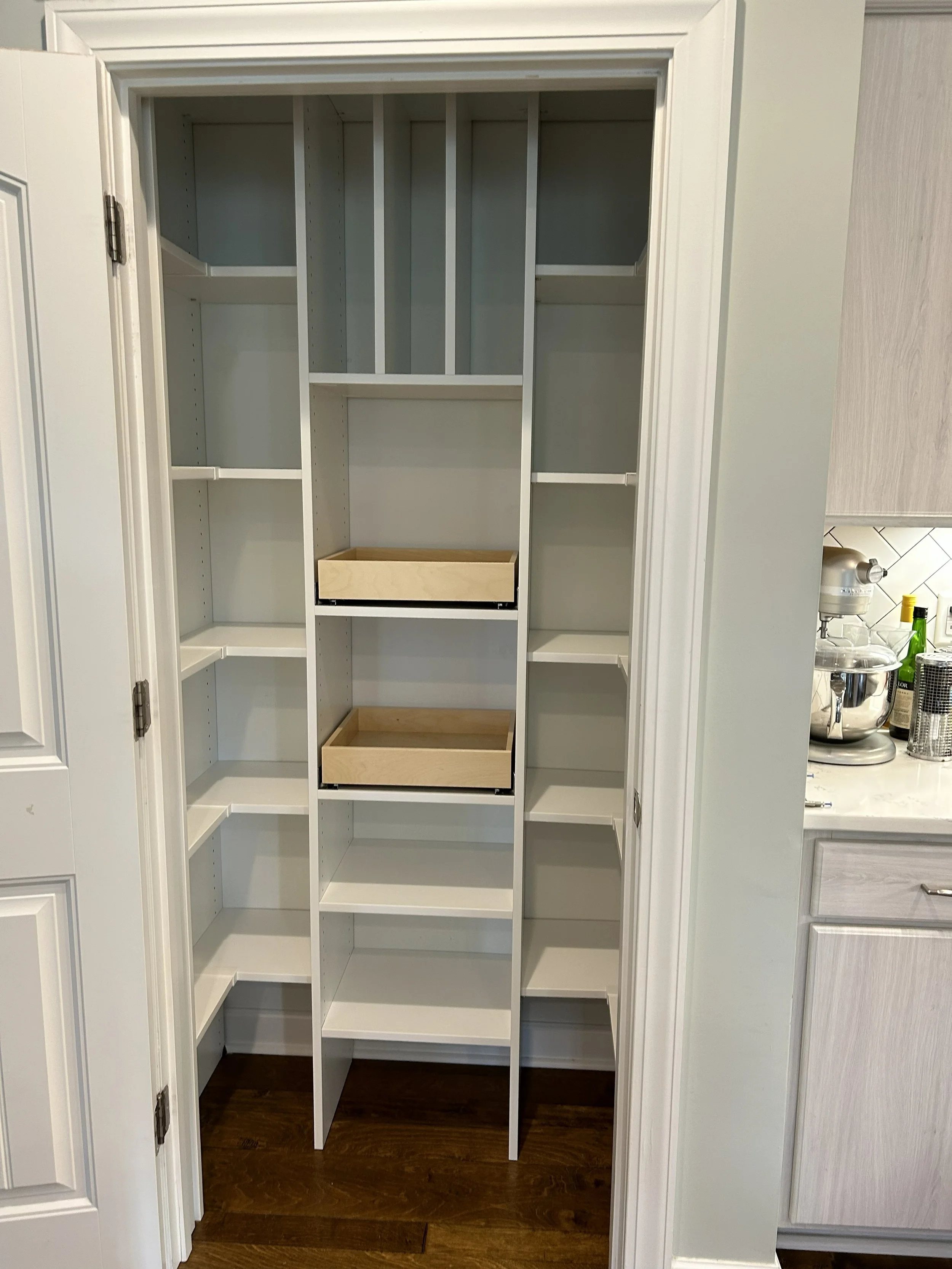 A custom kitchen pantry closet with multiple shelves, two wooden trays, and an open door showing a kitchen with counters, a mixer, and bottles.