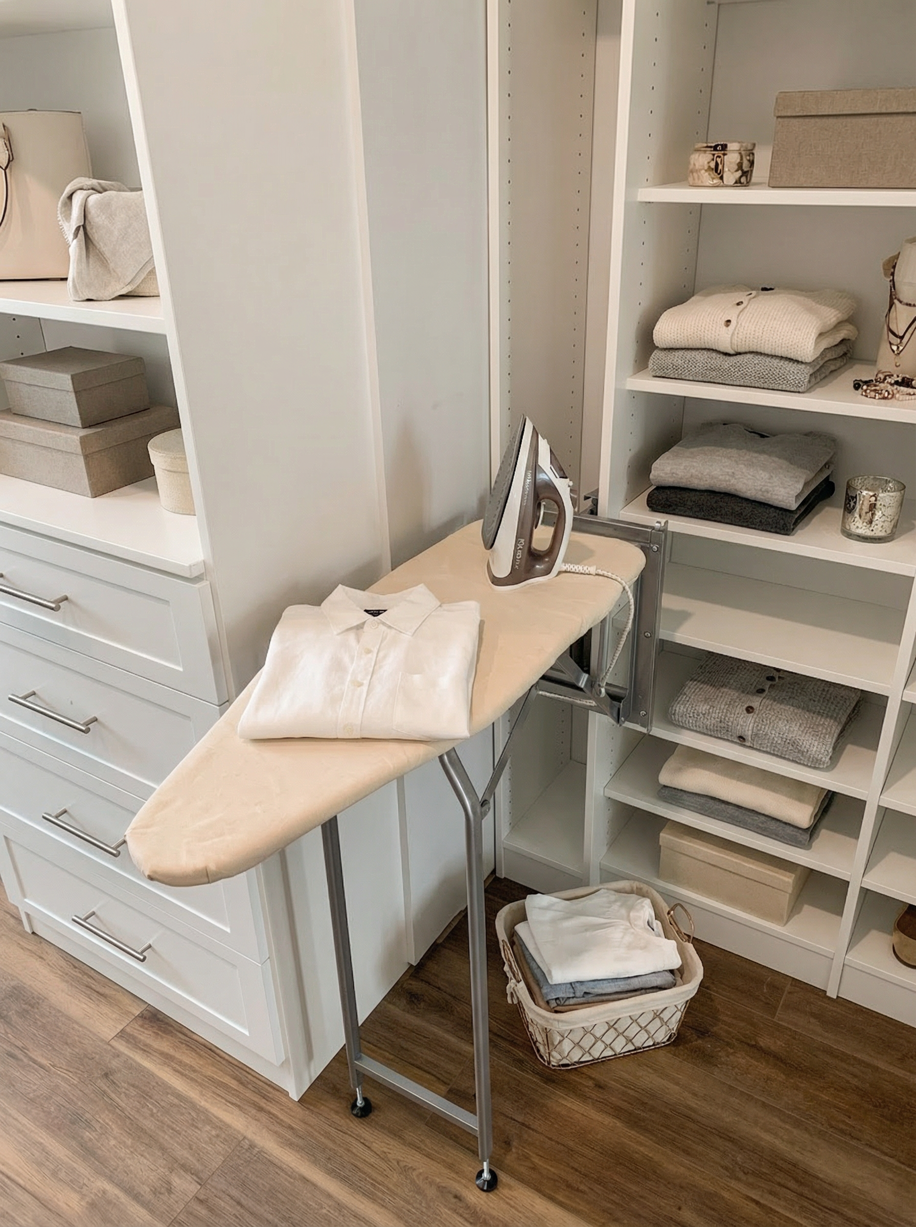 Clothes being ironed on an ironing board, with a folded shirt and an iron, in a walk-in closet with shelves of folded linens and storage boxes.