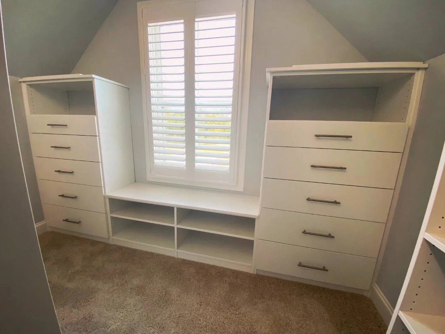 White built-in storage units with custom drawers and open shelves under a window with white shutters, in a room with beige carpet and light gray walls.