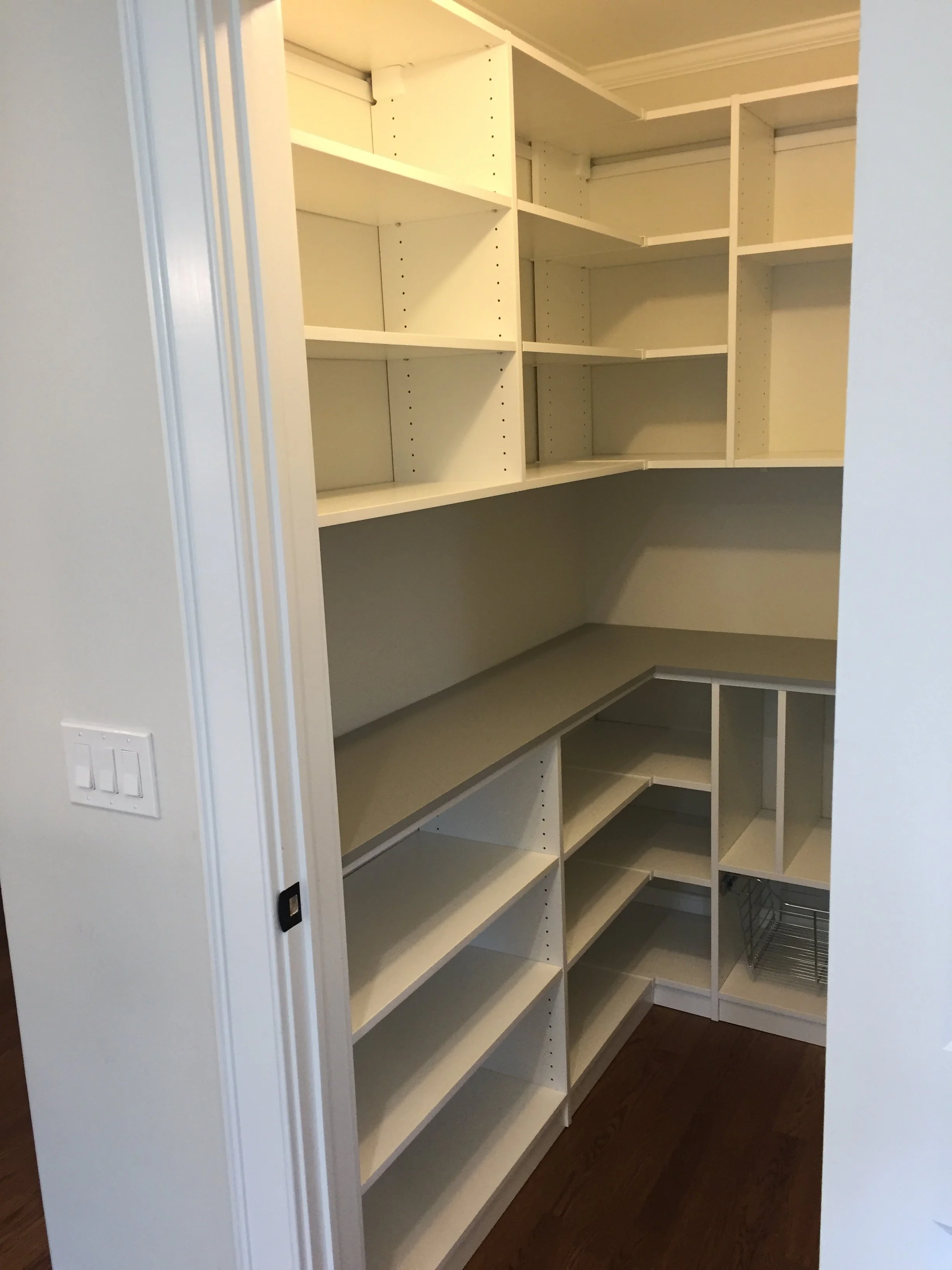 Built-in kitchen pantry with multiple shelves and a small wire basket, shown from an open door view on a hardwood floor.