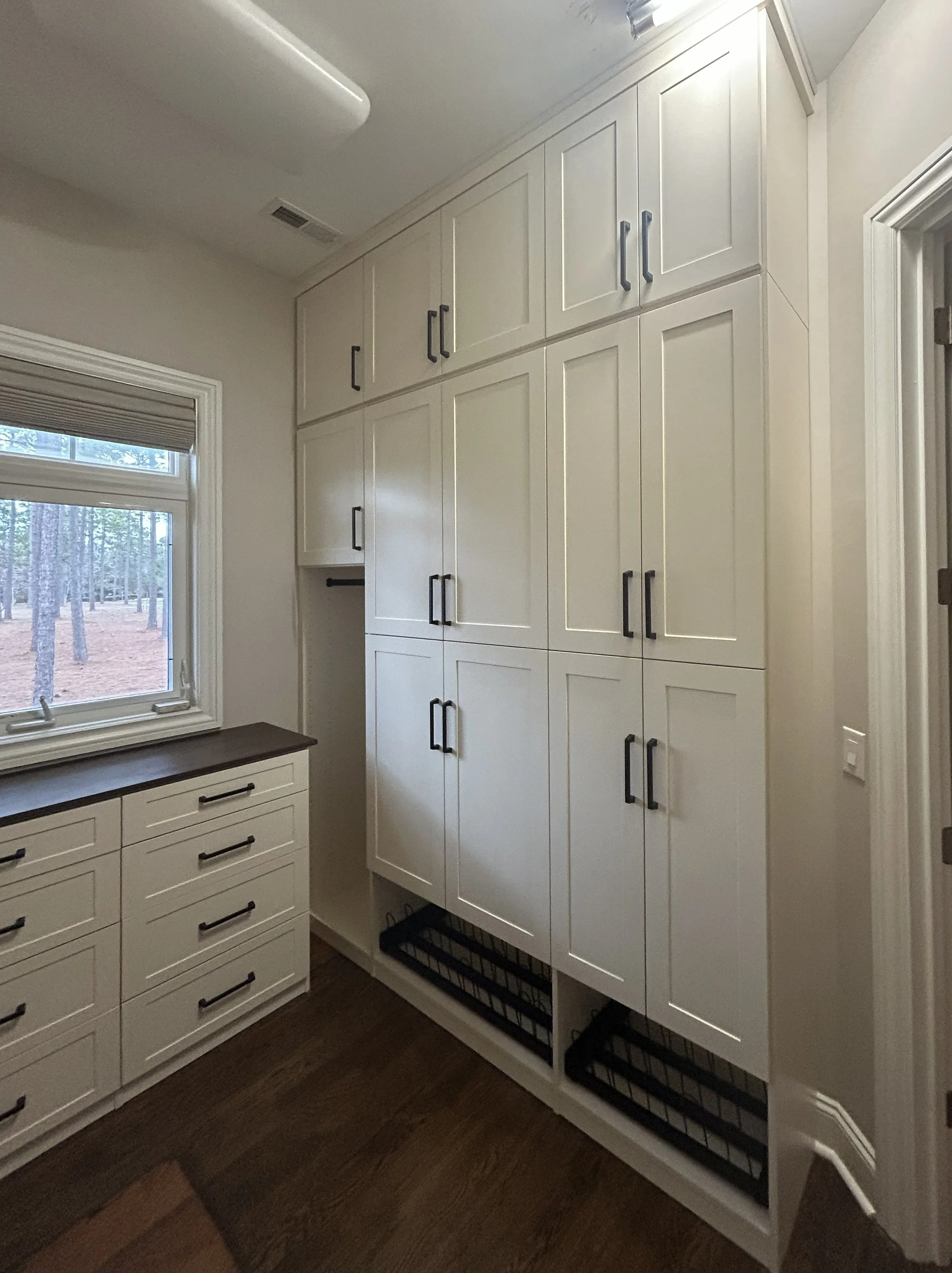 A room with custom white storage cabinets and drawers, window with a view of trees, and a dark countertop.