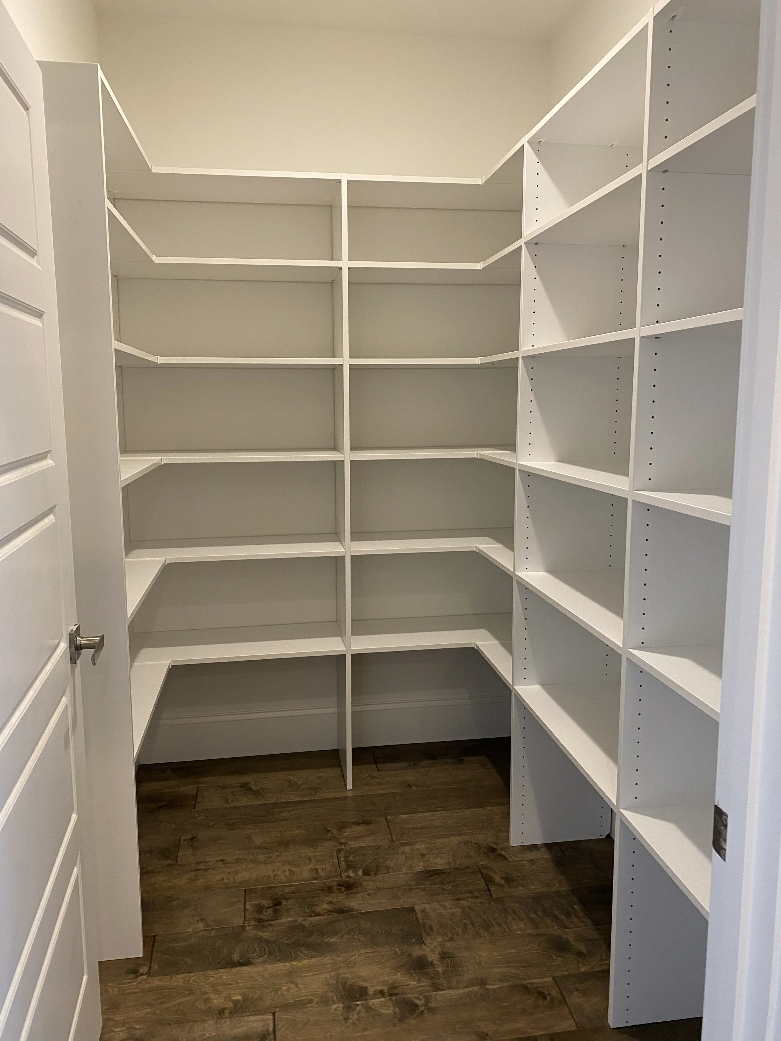 Walk-in kitchen pantry designed with white shelves and dark wood flooring.