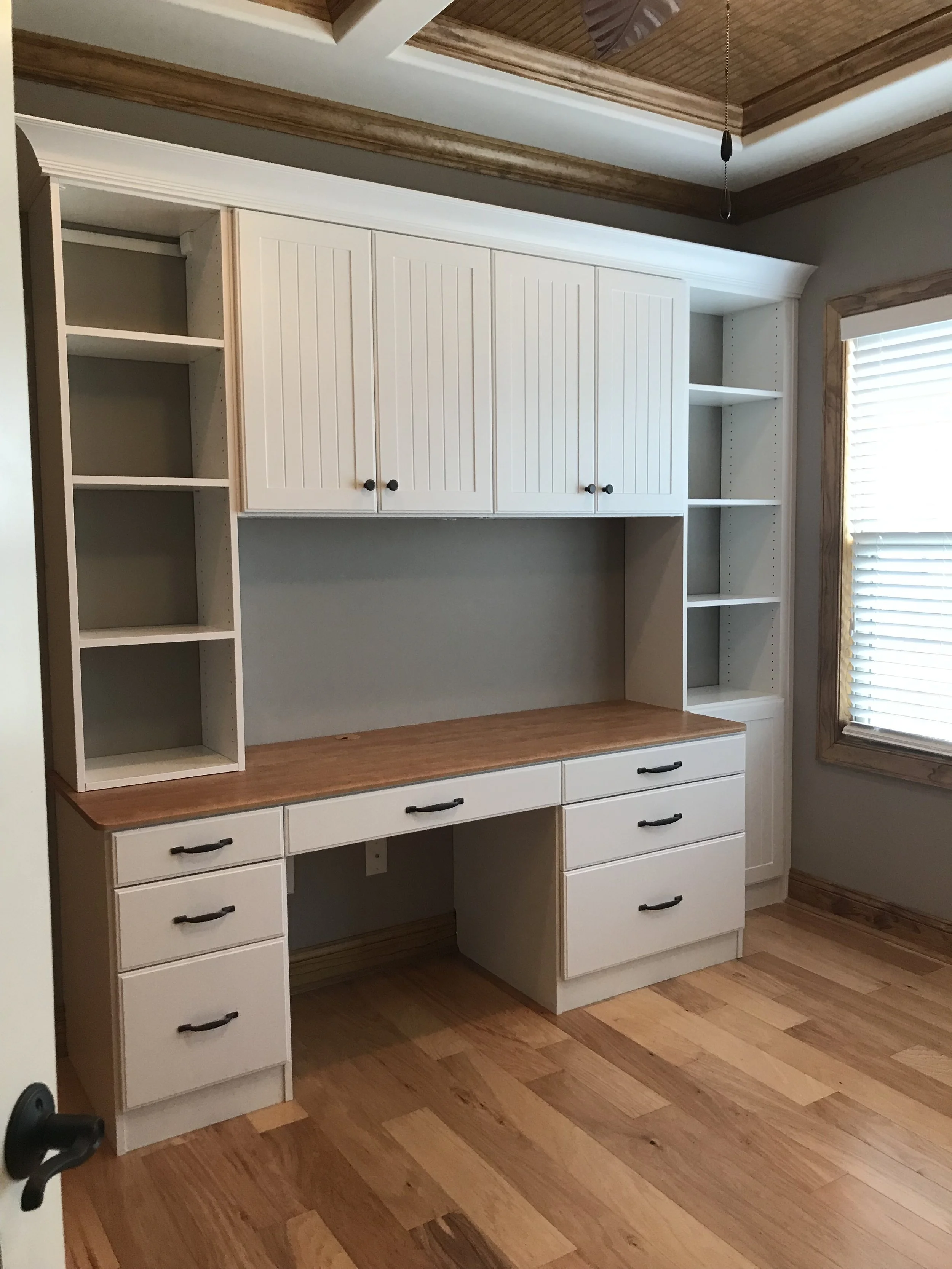 Built-in desk with office storage cabinets, drawers, and open shelves, situated near a window with blinds, in a room with wooden flooring and trim.