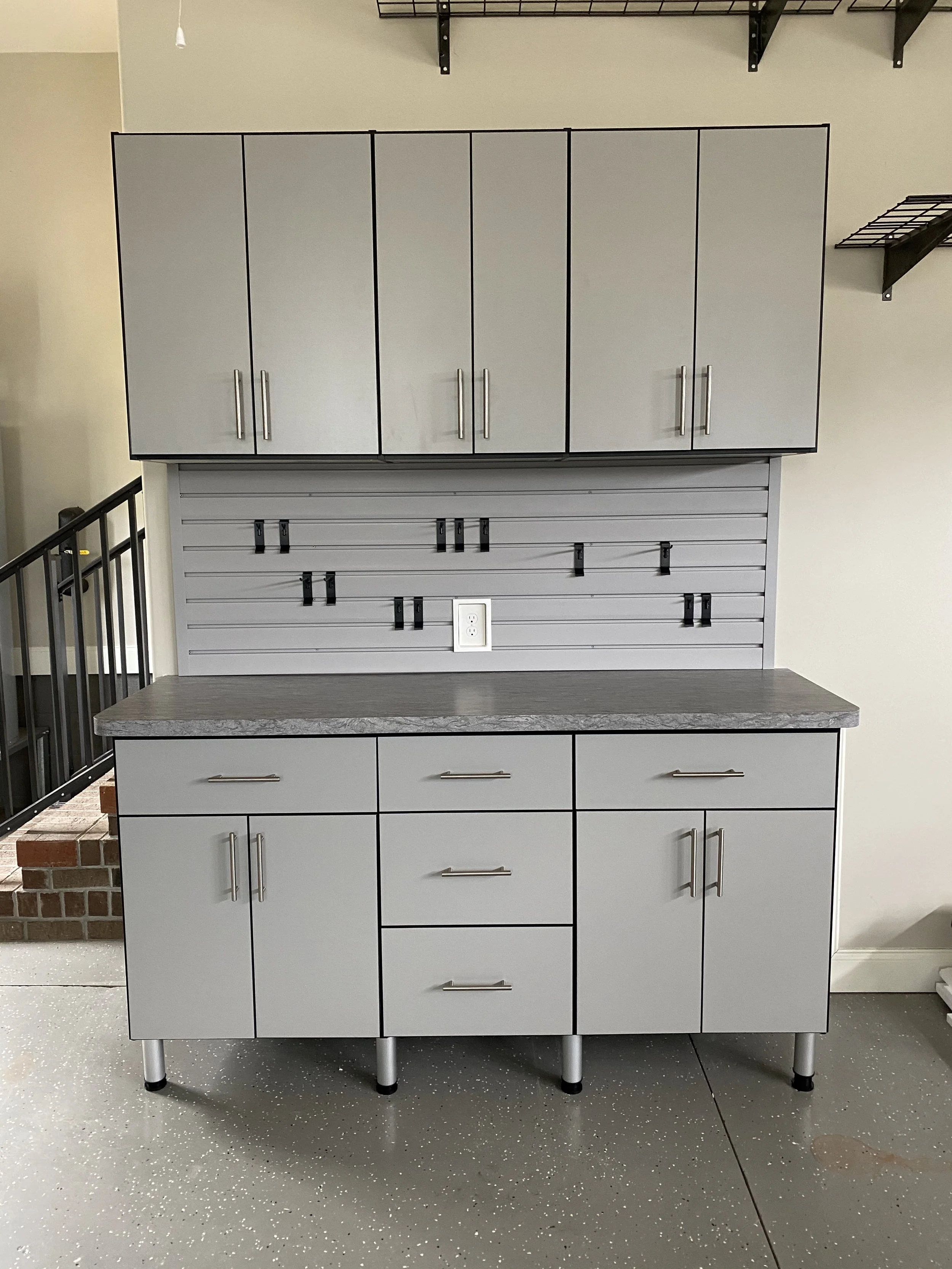 Gray grage  custom storage with upper and lower storage, a countertop, and a gray backsplash with hooks, in a room with a speckled gray floor.