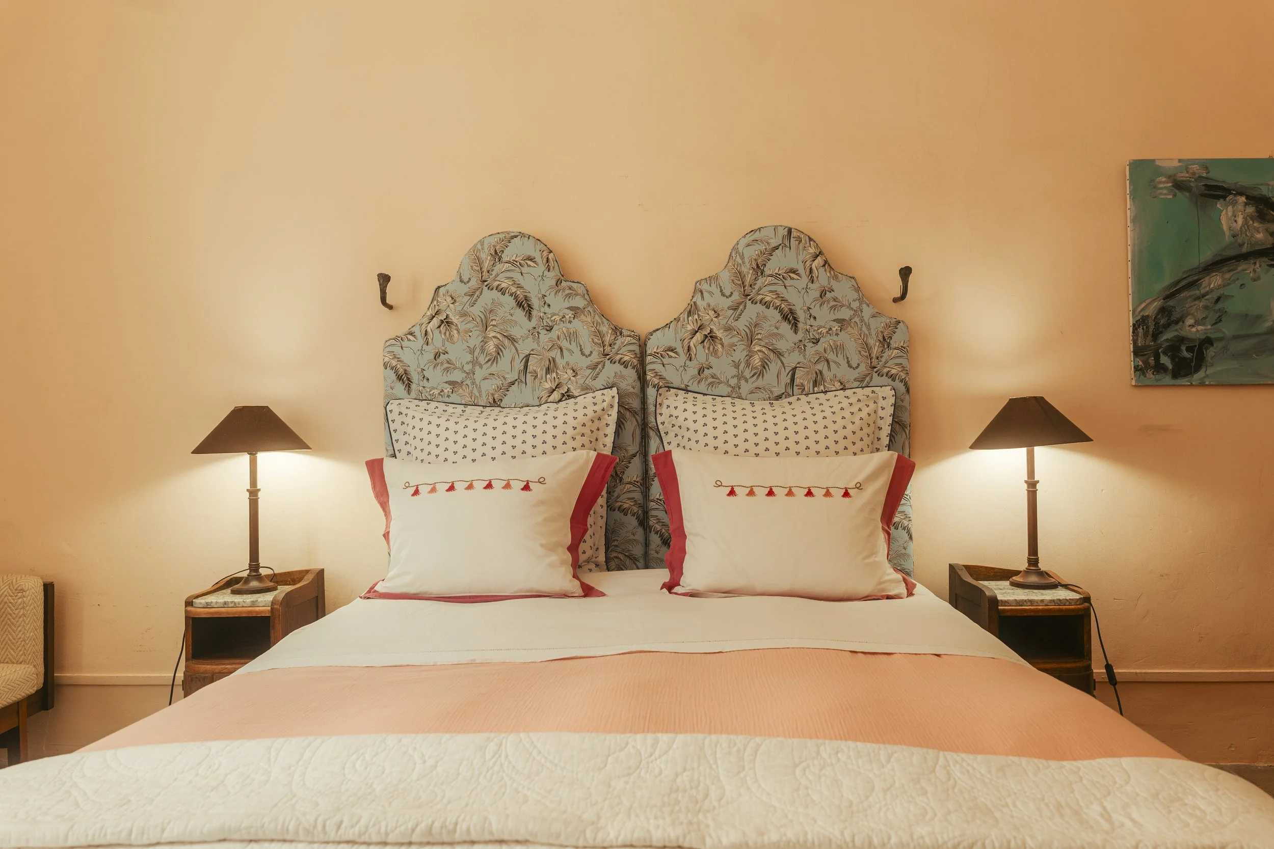 A neatly made bed with a floral patterned headboard, two pillows with white covers and red borders, and two table lamps on bedside tables, set against a beige wall with a painting hanging on the right side.
