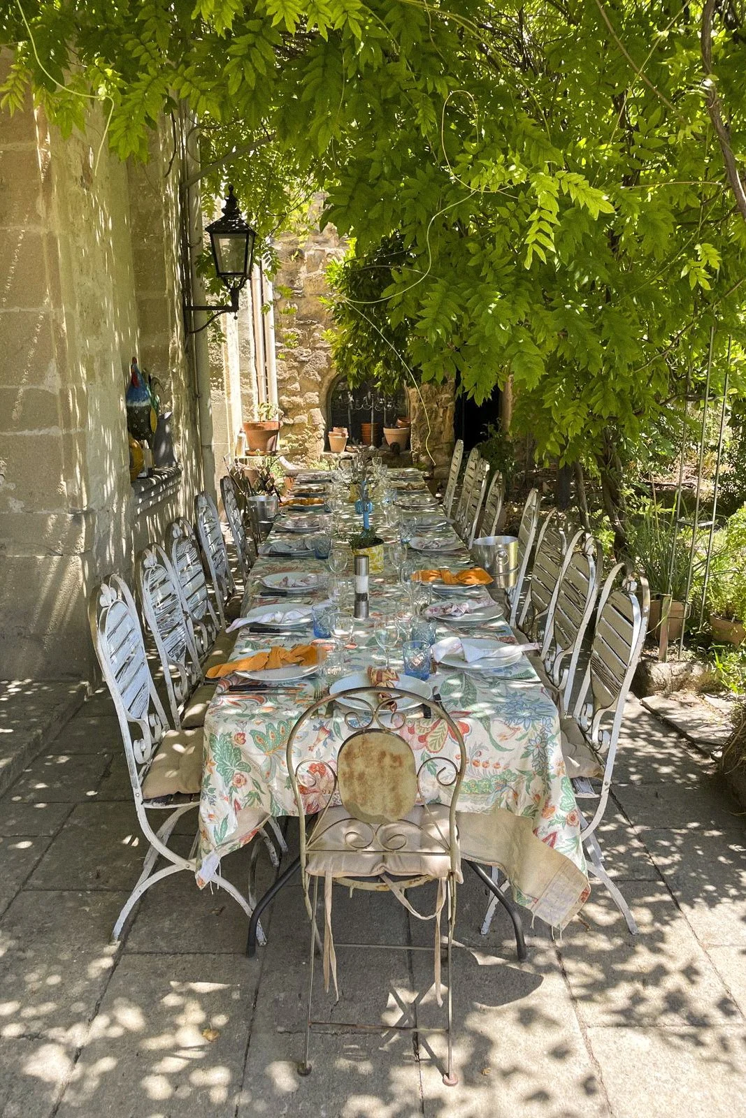 Long outdoor dining table set for a meal, surrounded by white chairs with floral tablecloths, under leafy green trees in a garden courtyard.
