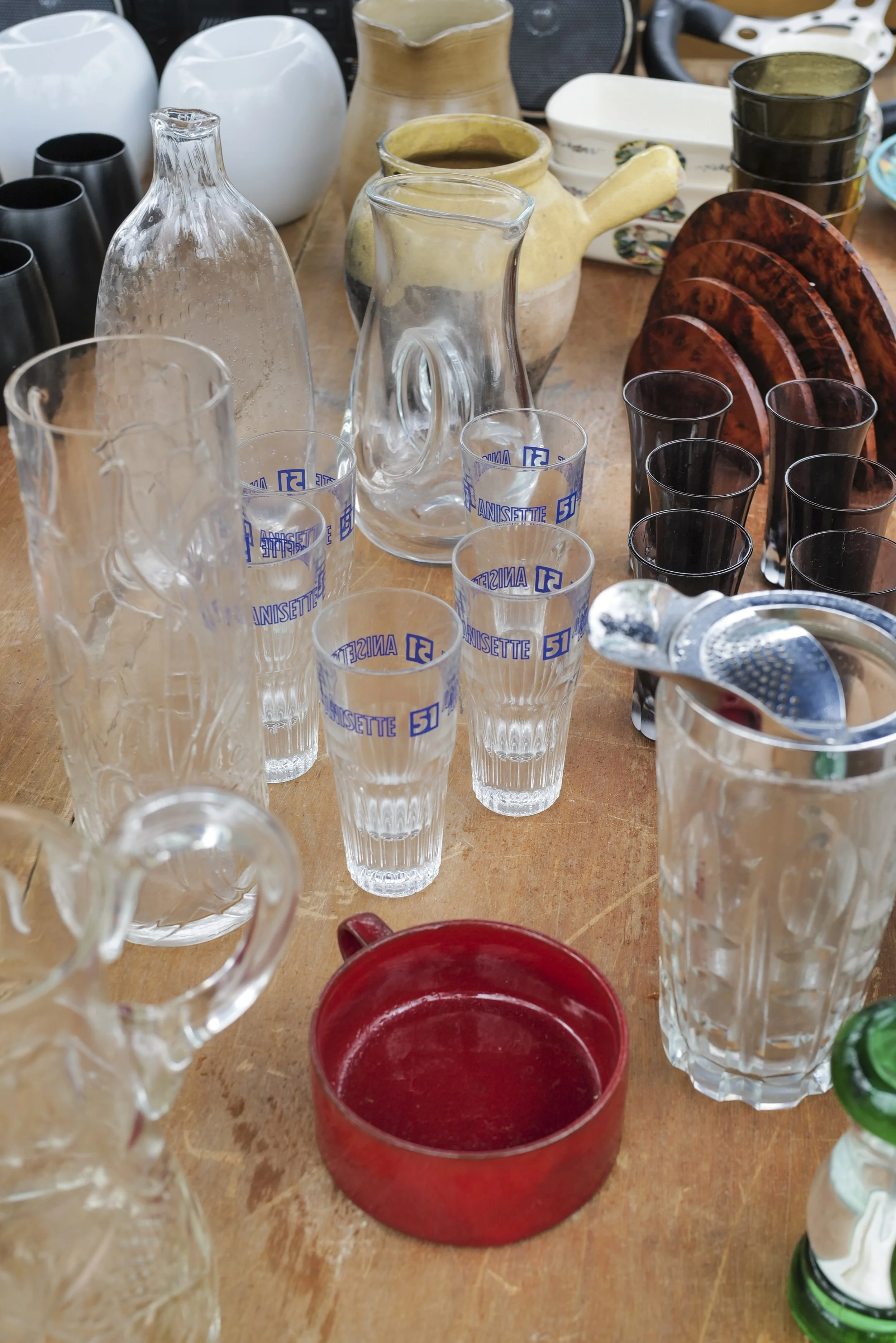 An assortment of glassware and ceramic items on a wooden table, including clear glass pitchers with handles, black and green glasses, a yellow ceramic teapot, and a red bowl.