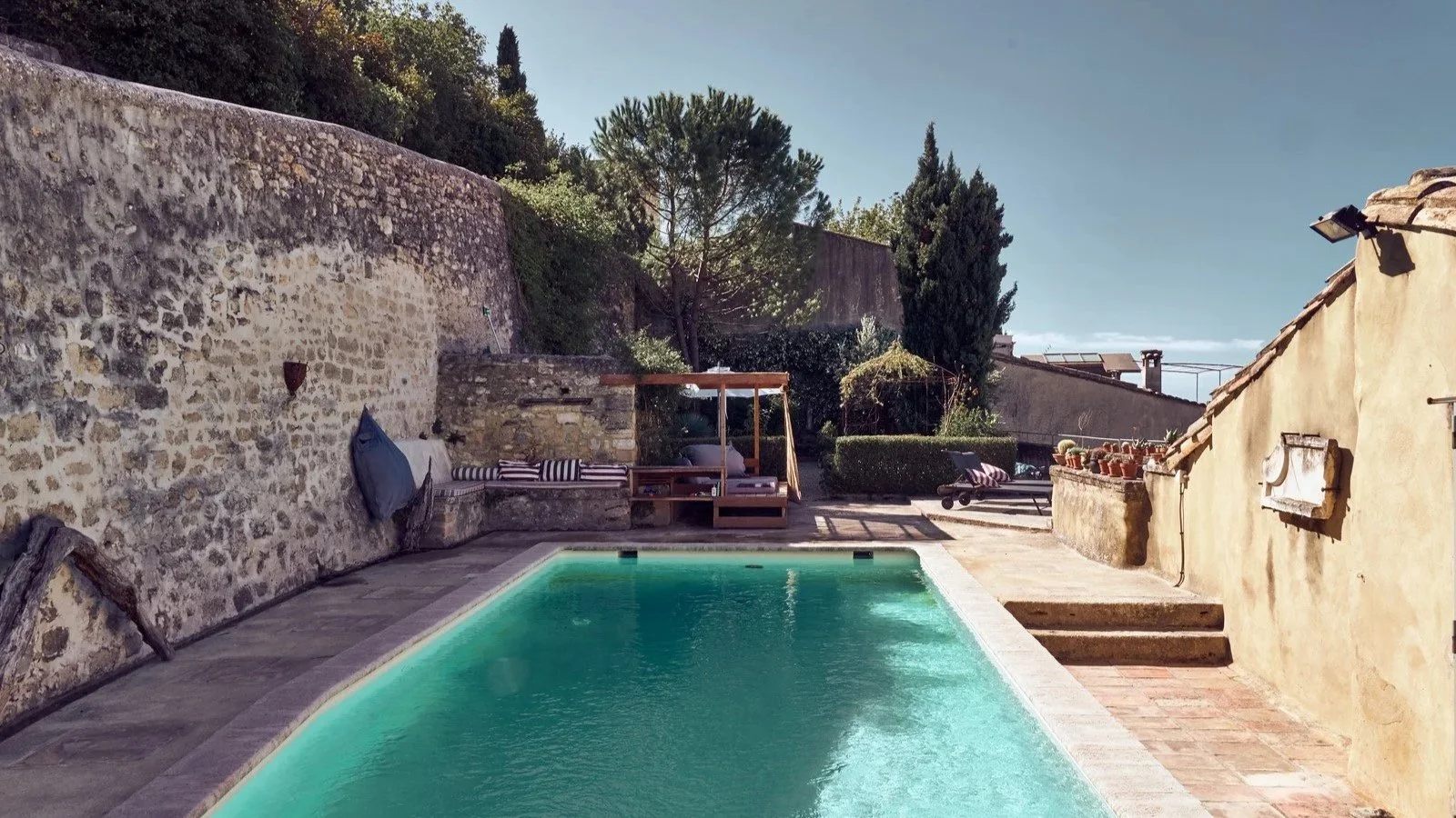 A backyard with a small swimming pool, stone and stucco walls, outdoor seating with striped cushions, and potted plants on a sunny day.