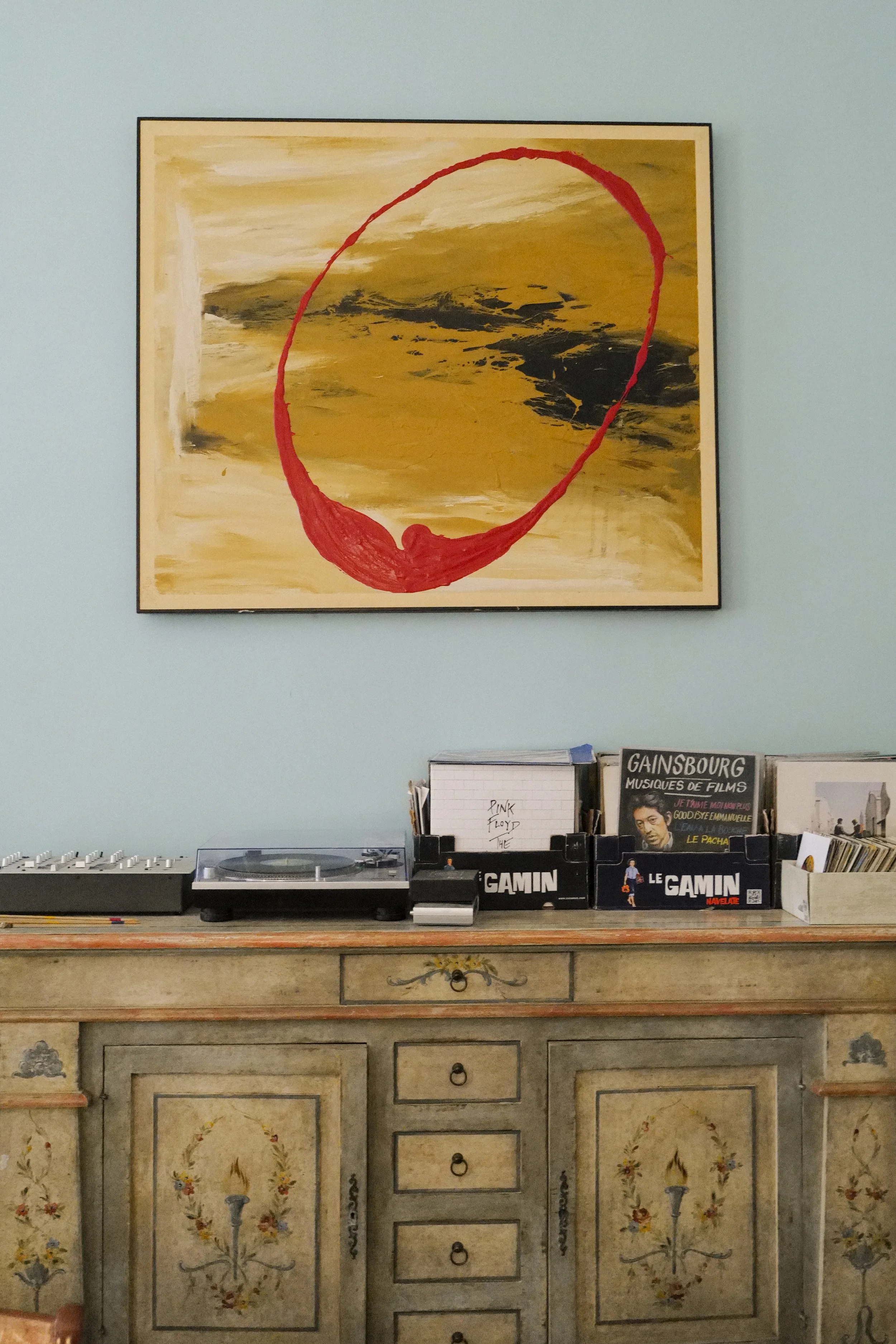 A vintage wooden sideboard with floral painted designs, a modern turntable, and record collection, against a pastel blue wall. Above is abstract artwork with bold brushstrokes and a red circular shape.