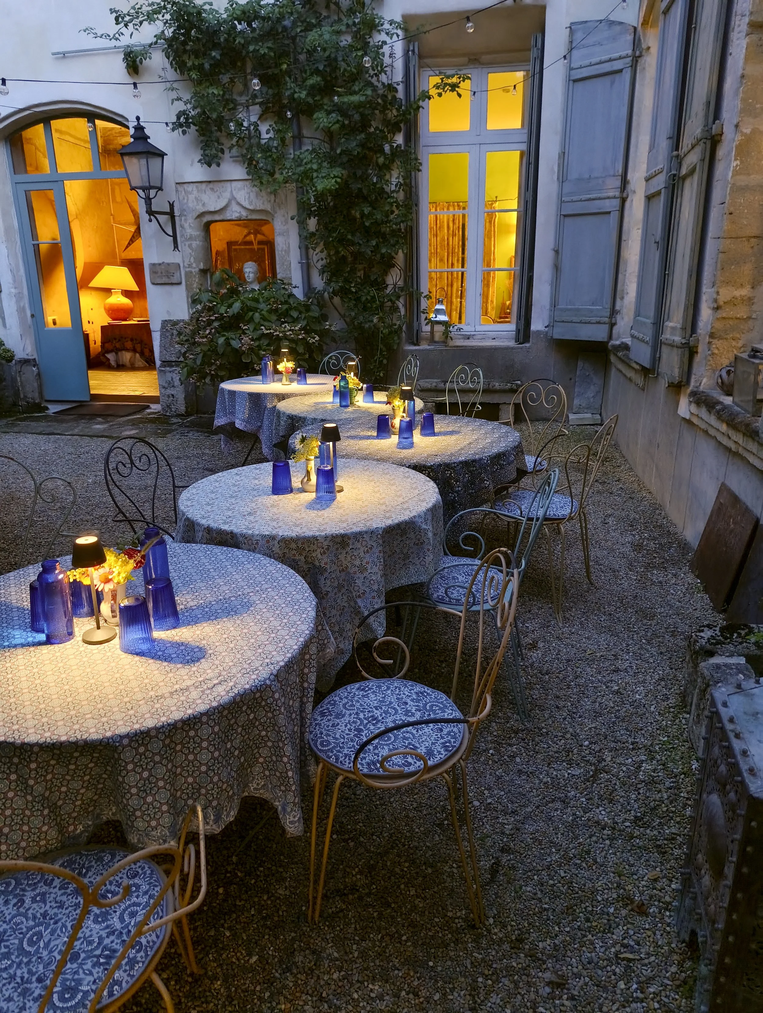 An outdoor dining area at dusk with round tables covered in blue and white patterned tablecloths. Each table has blue cups, small vases with yellow flowers, and mini black lamps. The background shows a building with light blue shutters, a window with