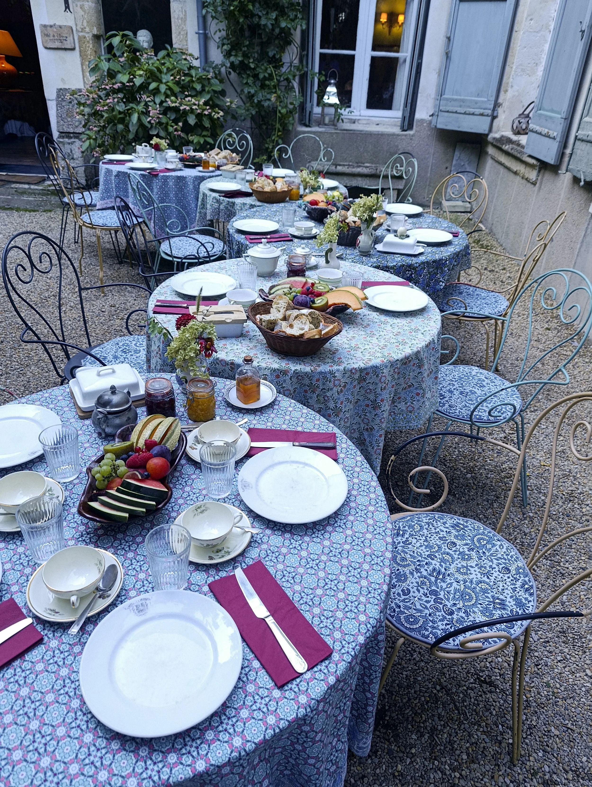 Outdoor dining table set for breakfast with plates, cups, utensils, fresh fruits, bread, honey, jams, and floral centerpieces on a patterned tablecloth in a cozy courtyard with open window shutters.