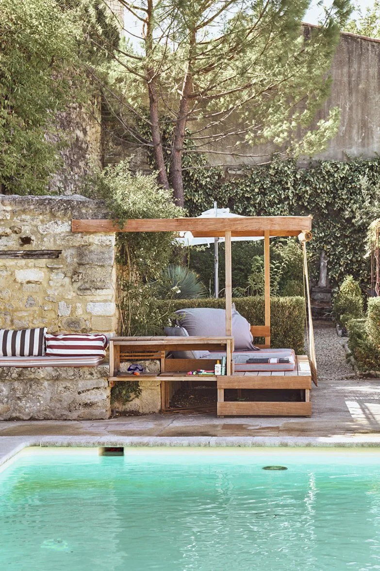 A backyard scene with a swimming pool in the foreground, and a wooden bench with cushions and a canopy in the background surrounded by greenery and trees.