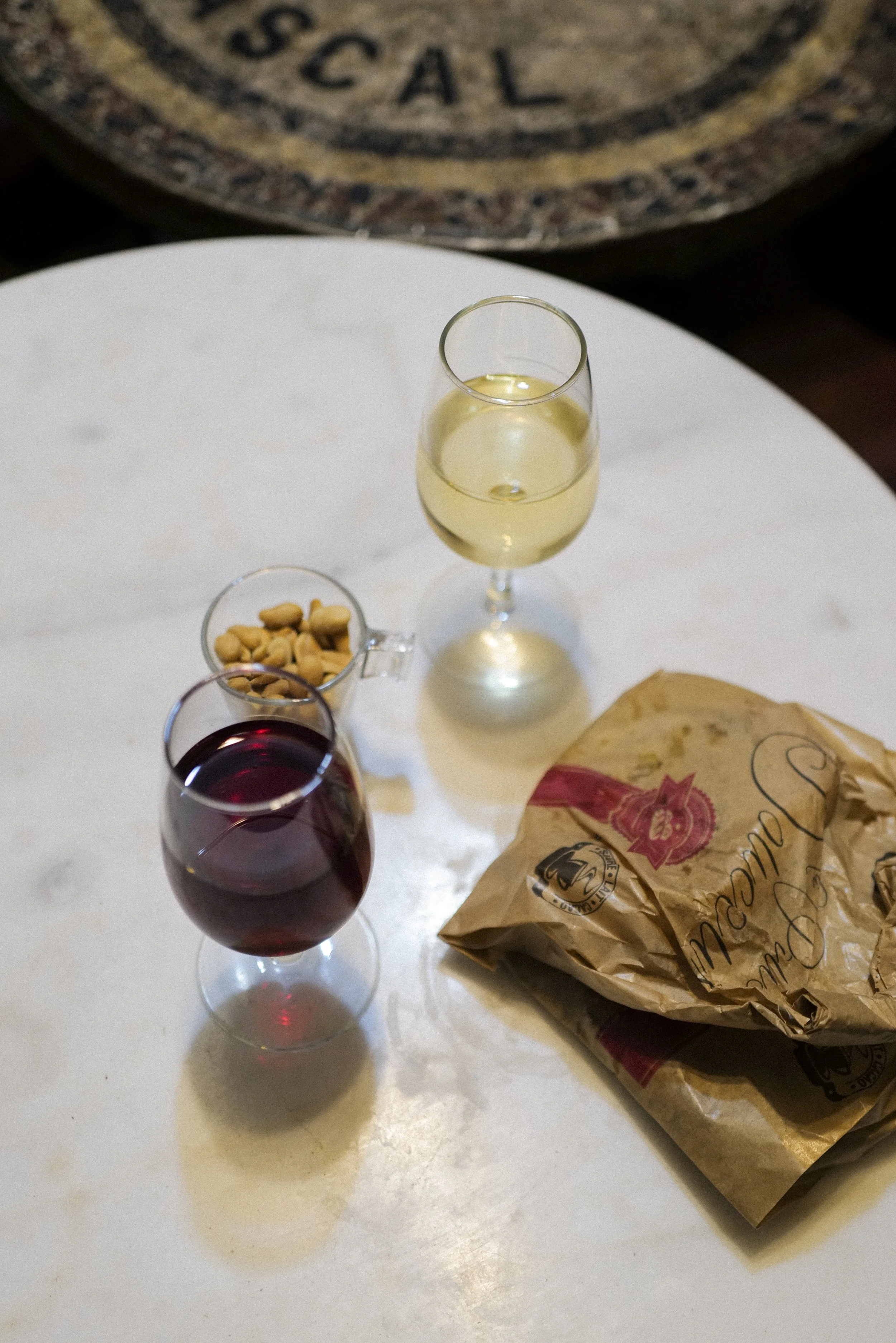 A table with two glasses of red wine, one glass of white wine, a small bowl of nuts, and a brown paper bag of bread, with a blurred decorative object in the background.