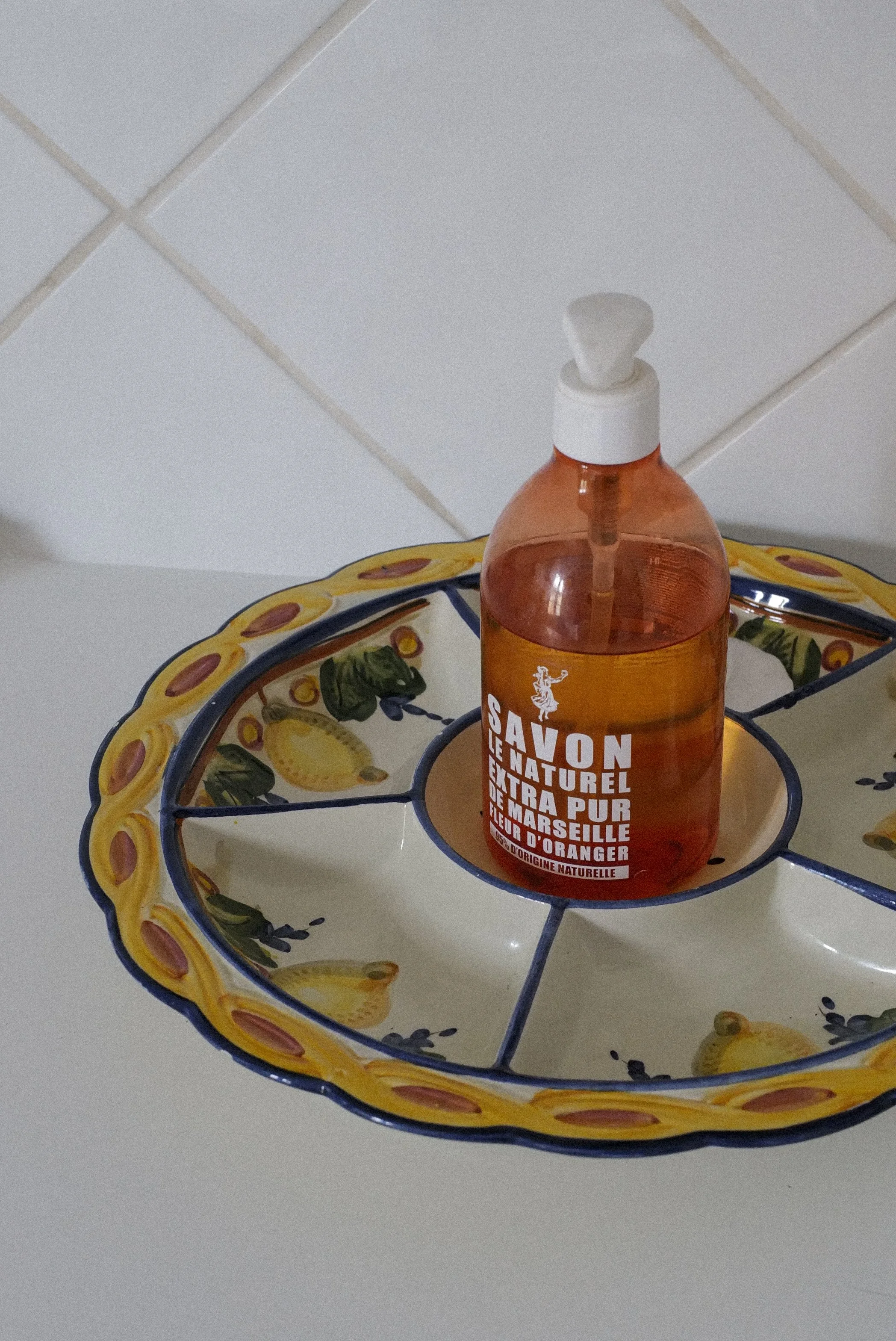 A bottle of Savon naturel extra pur, with a white cap and orange soap inside, placed on a decorative ceramic tray with a blue and yellow pattern.