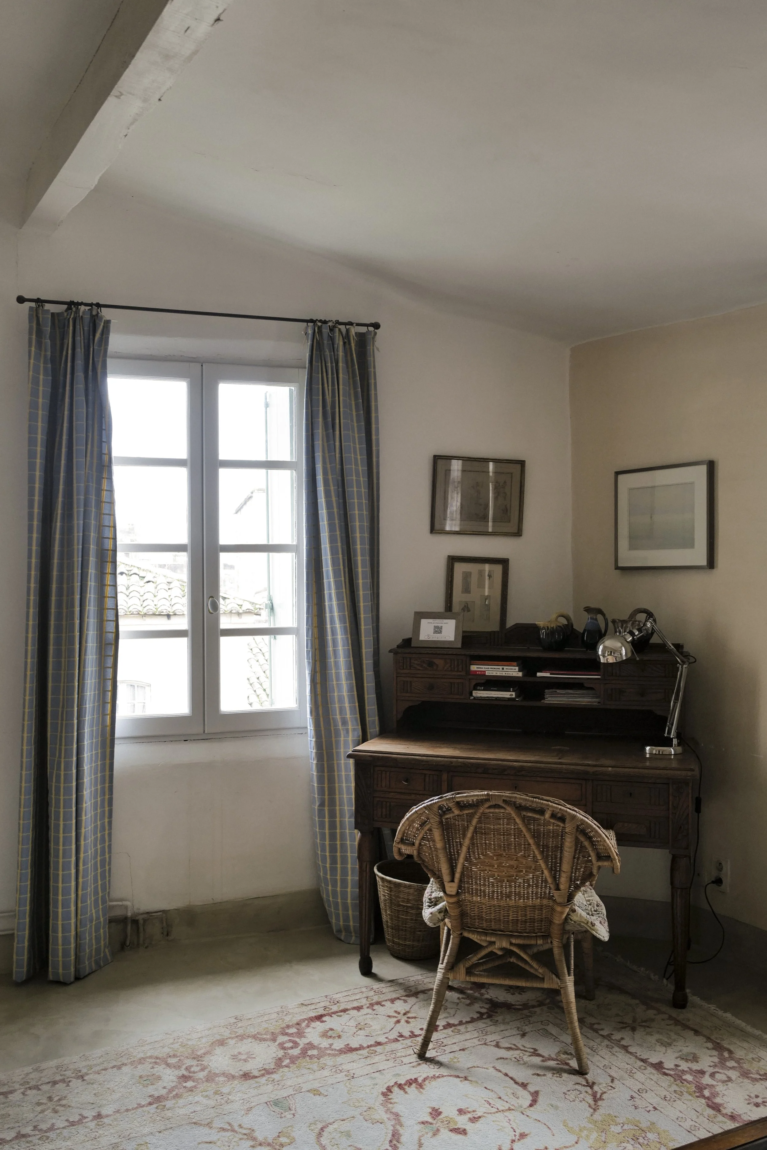 A study or sitting area with a window, blue plaid curtains, a wooden writing desk with framed pictures on it, a wicker chair, a lamp, and a patterned rug.