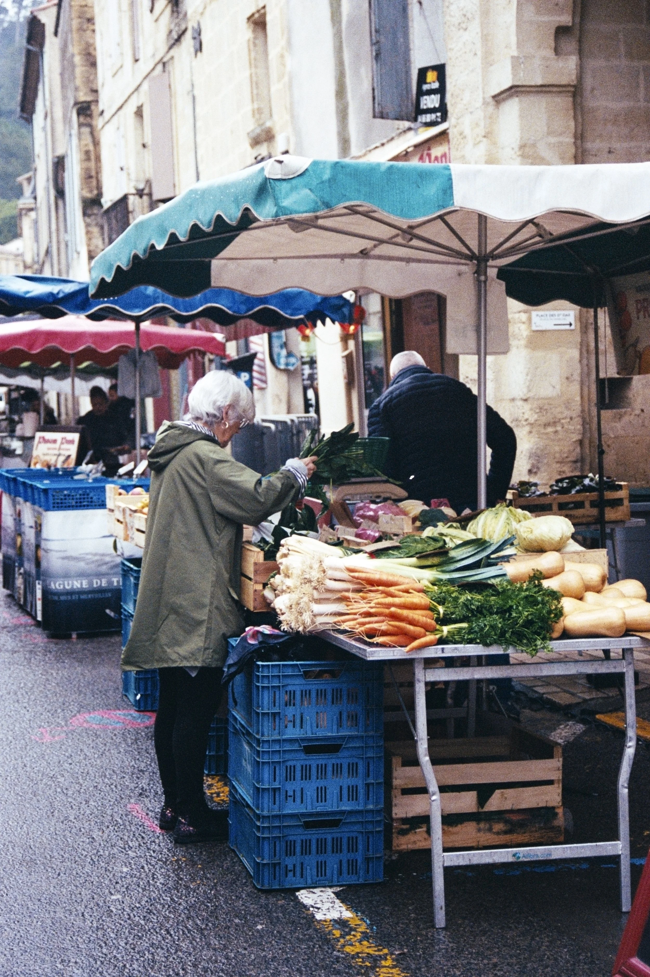 A street market scene with vegetables on display, including carrots, fennel, and green leafy vegetables, under umbrellas. A woman with white hair and glasses is shopping, and a man is behind the stall. The street has old stone buildings and a sign in