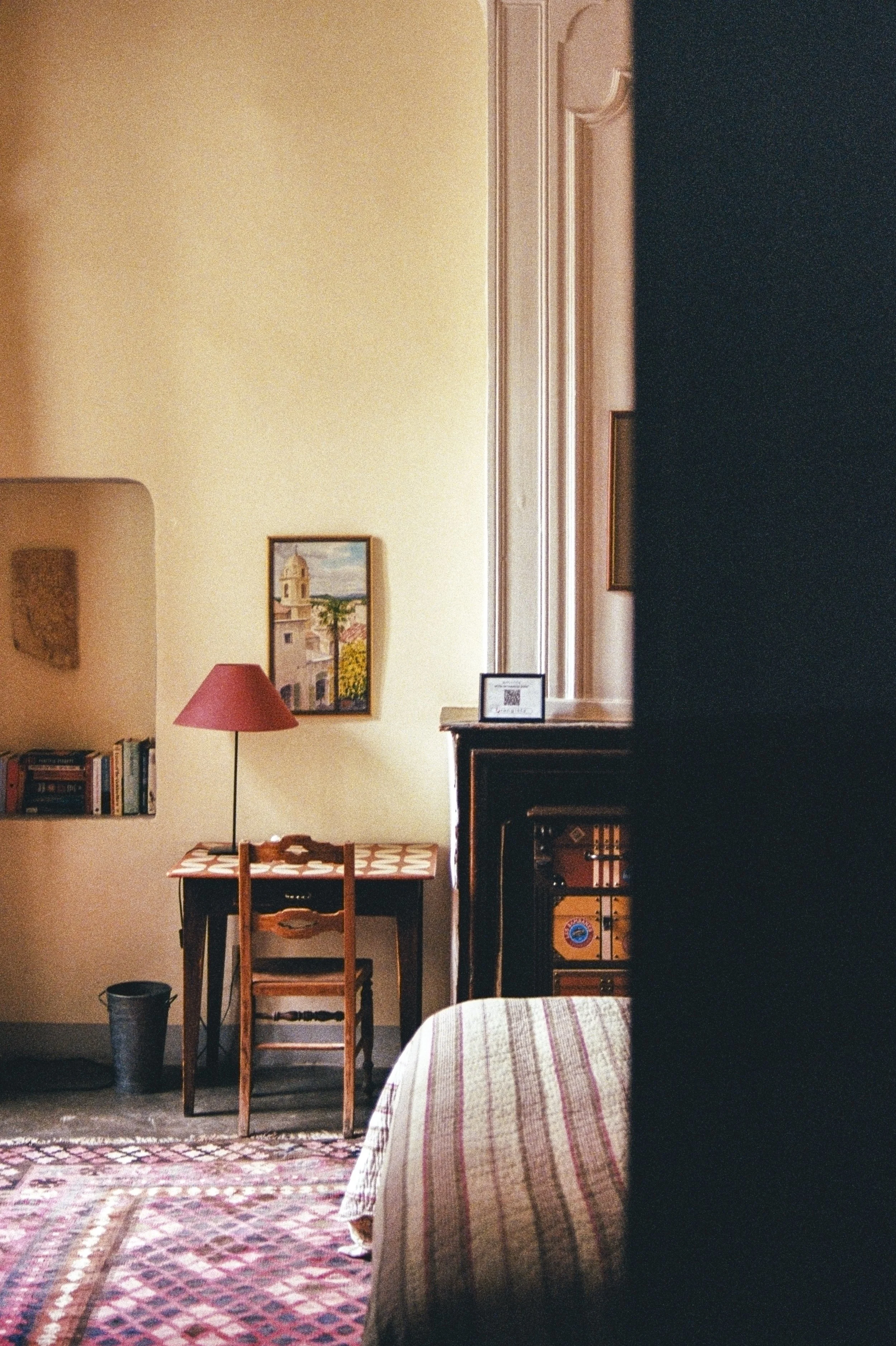 Interior view of a cozy room with warm lighting, featuring a painting of a building with a bell tower on the wall, a red table lamp, a small bookshelf, a wooden chair at a table with a checkered tablecloth, a patterned rug, a black wastebasket, and a